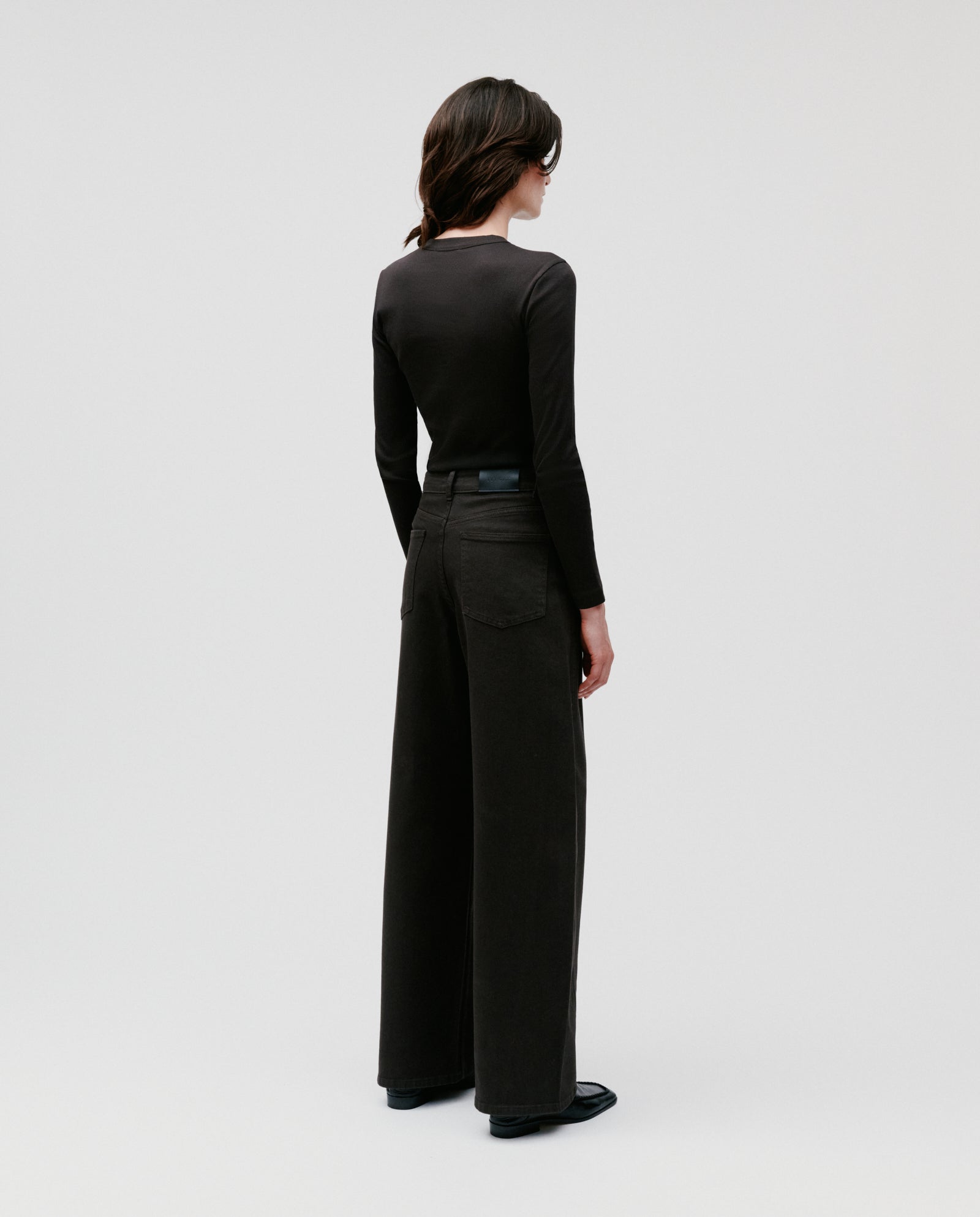 A woman in IVY OAKs PHILA Pants and a black long-sleeve top stands facing away against a plain light background.