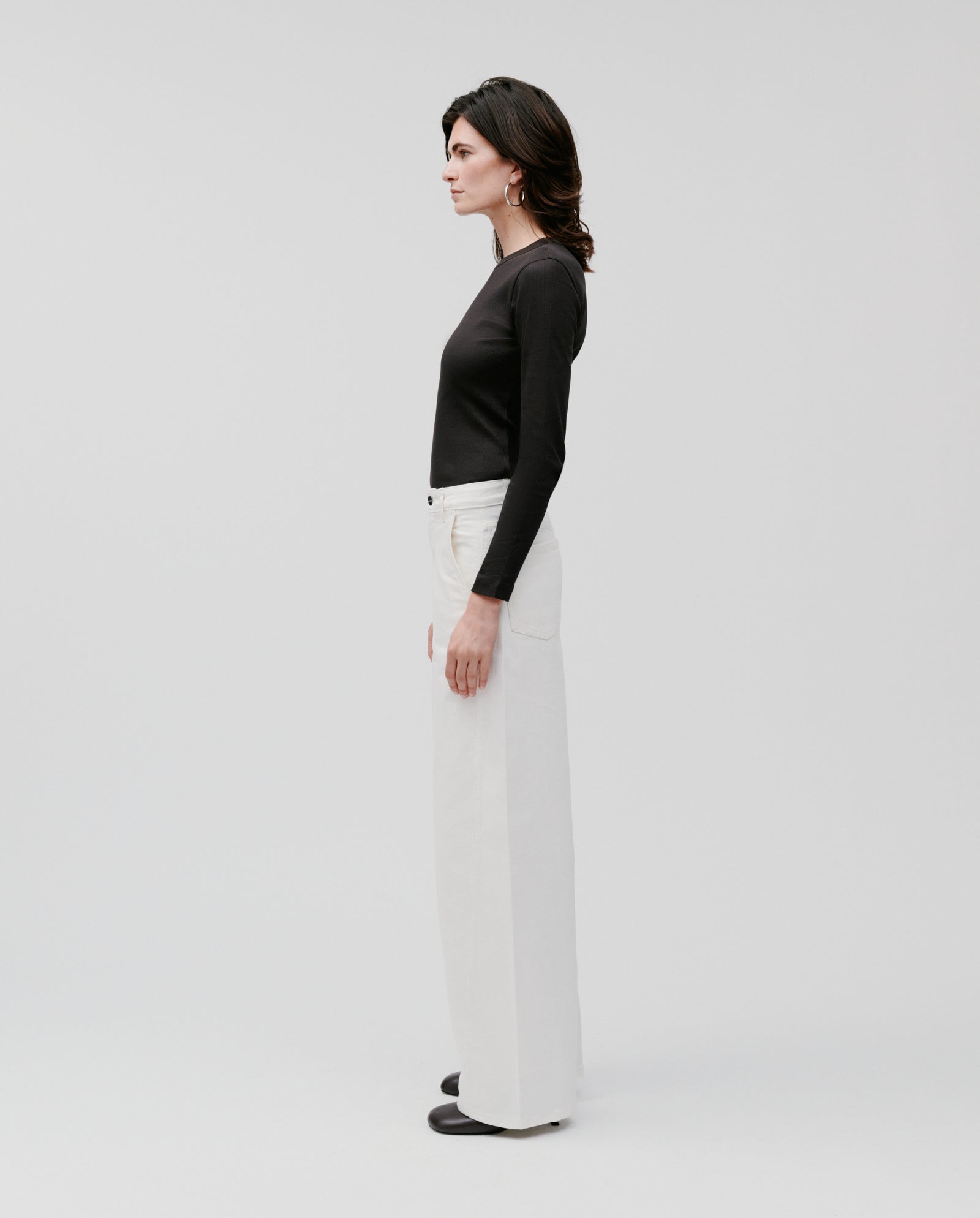 A woman stands in profile against a plain light background, wearing a black long-sleeve top and IVY OAKs PHILA Pants in a white barrel leg style.
