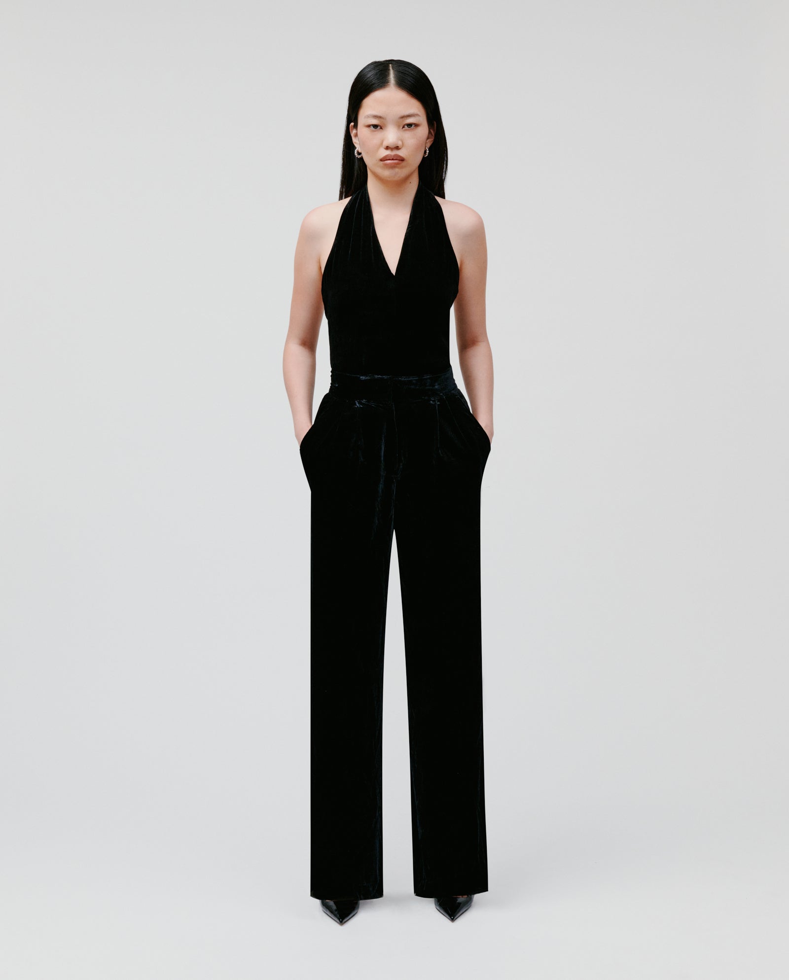 A woman wearing PRESCILLIA ANN Pants by IVY OAK—a black halter-neck jumpsuit with wide-leg velvet pants—stands with hands in pockets against a plain light background.