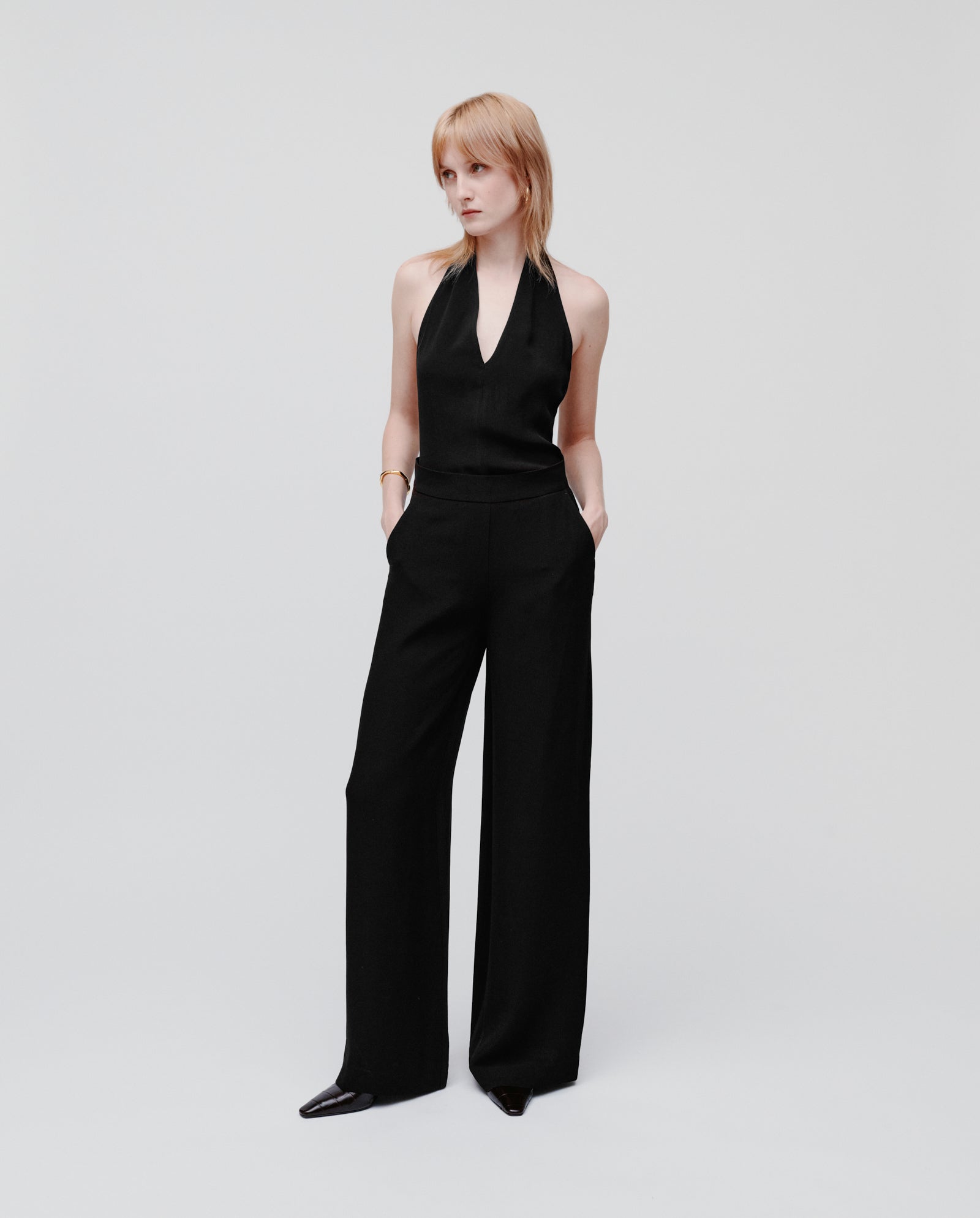 A person is wearing IVY OAK’s PHILINE SUE Pants—a minimalistic, wide-leg black trouser—styled with hands in pockets.