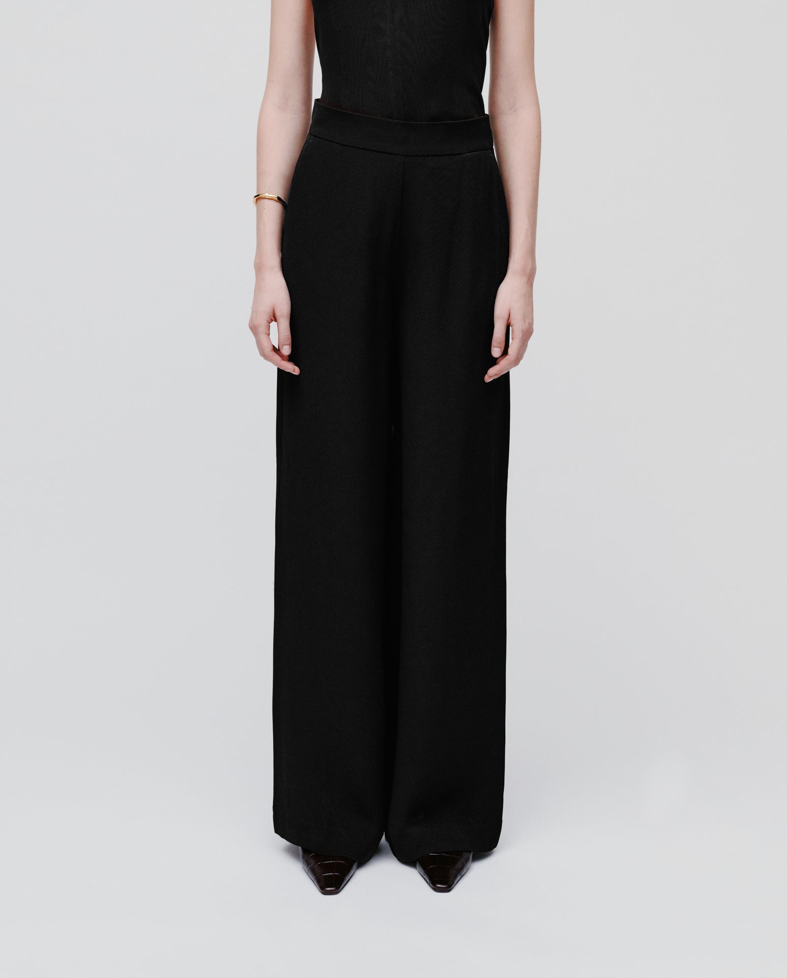 A person wears IVY OAKs PHILINE SUE Pants—minimalistic wide-leg trousers in black crepe fabric—with a sleeveless top, standing against a plain light background.