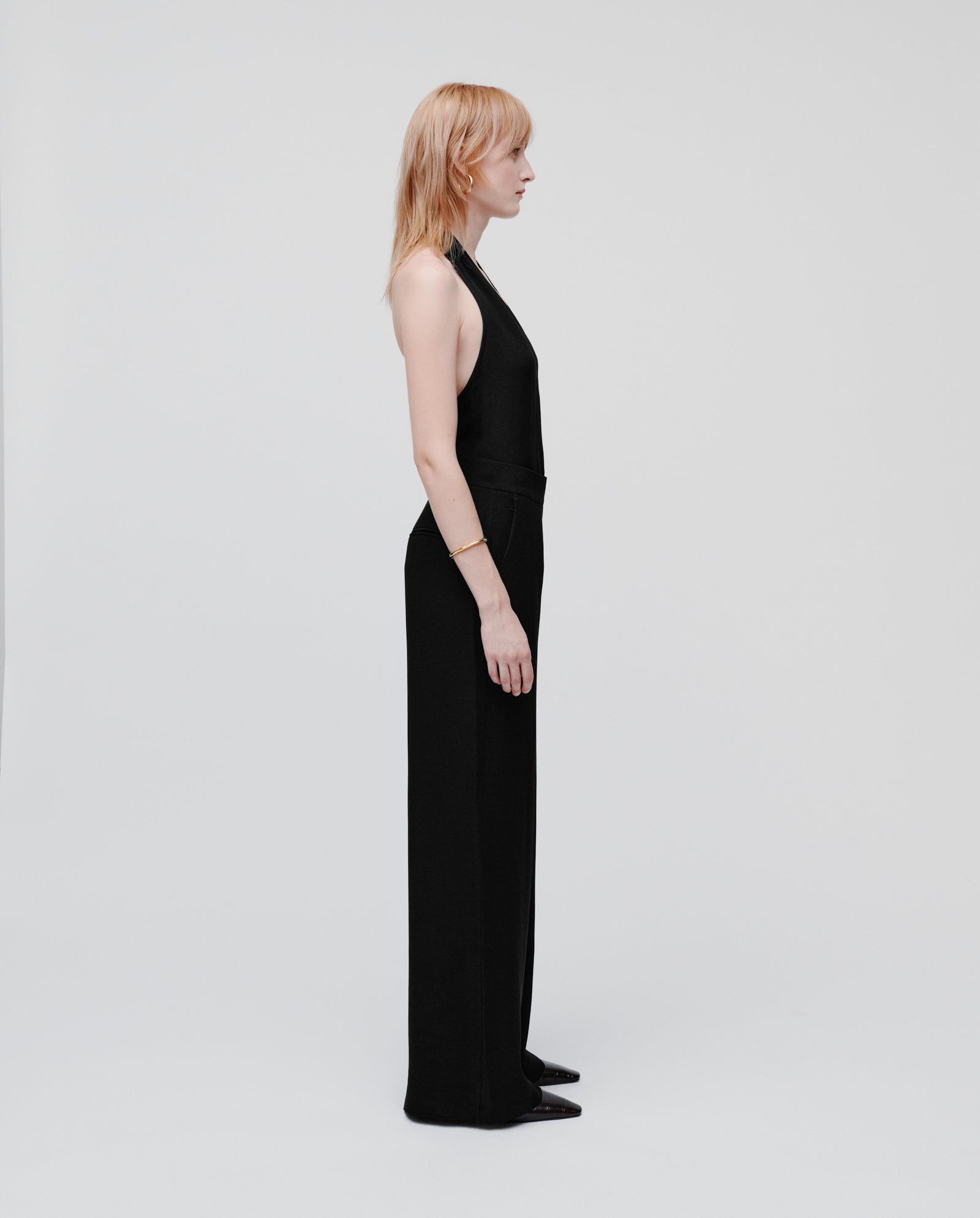 A person with light hair stands in profile against a plain white background, wearing the black IVY OAK PHILINE SUE Pants, a sleeveless jumpsuit with wide leg trousers.