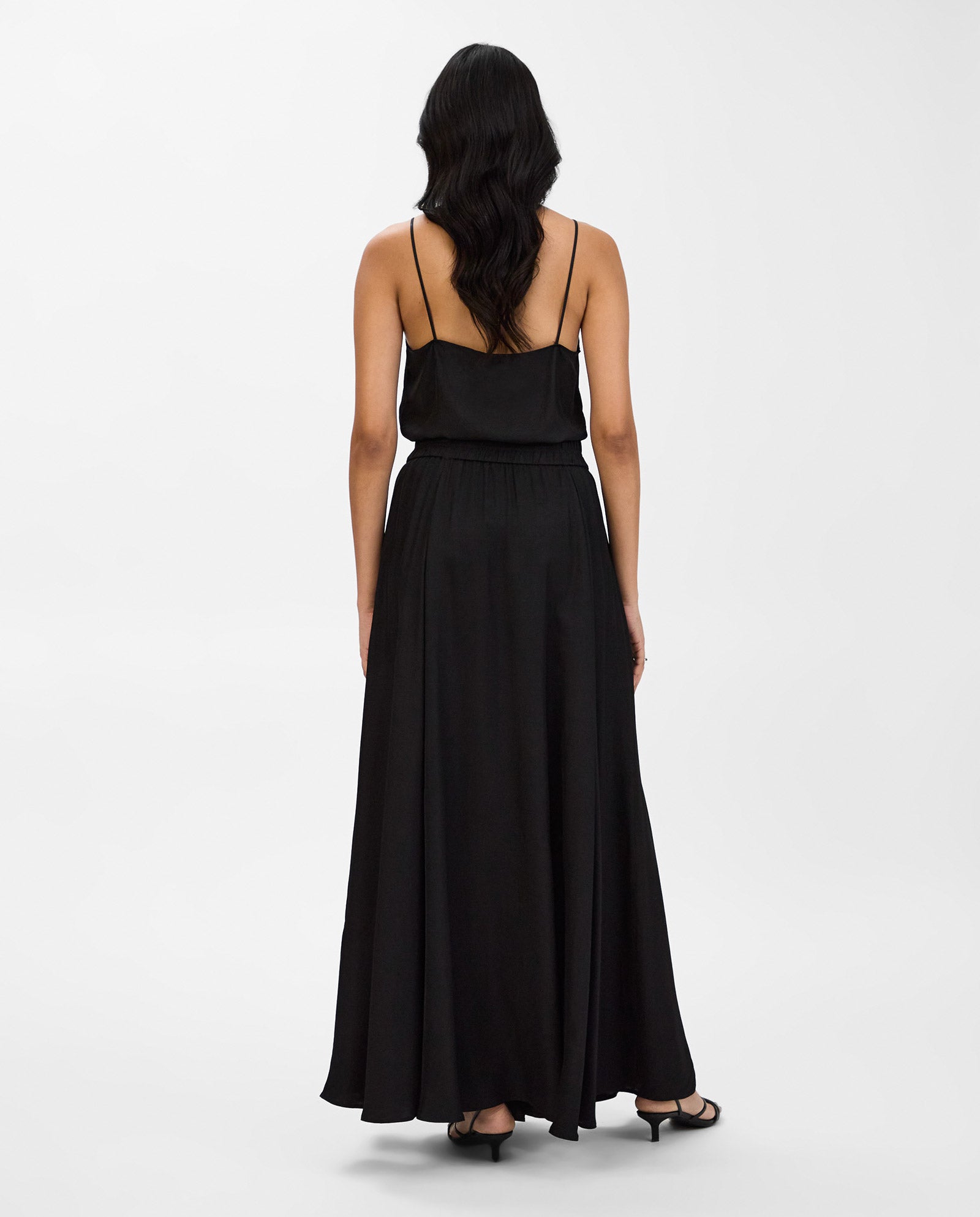 A woman wears the IVY OAK SYLVIA MAY Skirt, featuring a long black design with thin straps and an elegant maxi skirt, standing against a plain white background.