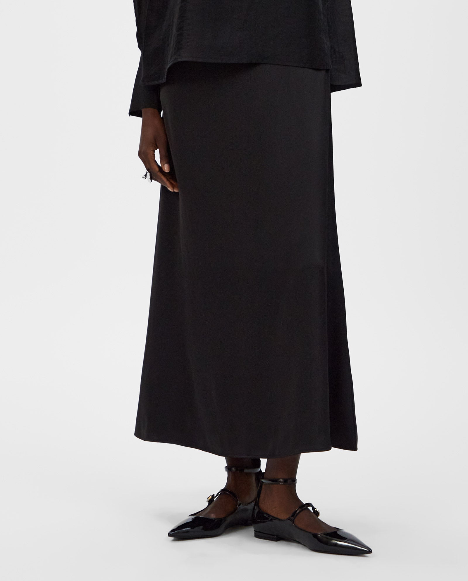 A person wears an ankle-length black IVY OAK SUMMER Skirt in satin, paired with black shoes, standing against a plain white background.