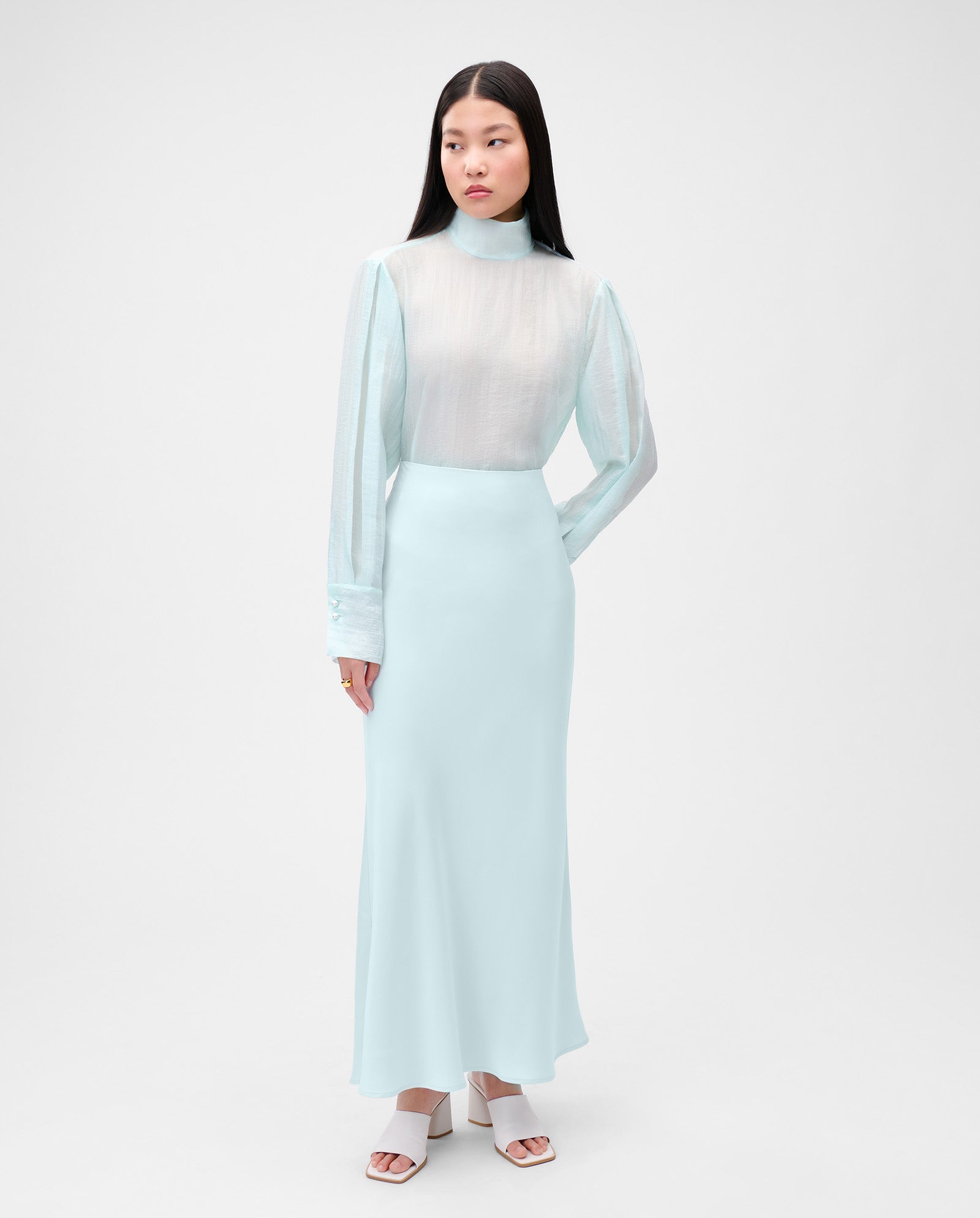 A woman wears IVY OAK’s SUMMER Skirt—a pale blue satin slip skirt—styled with a sheer white long-sleeve top and white sandals.
