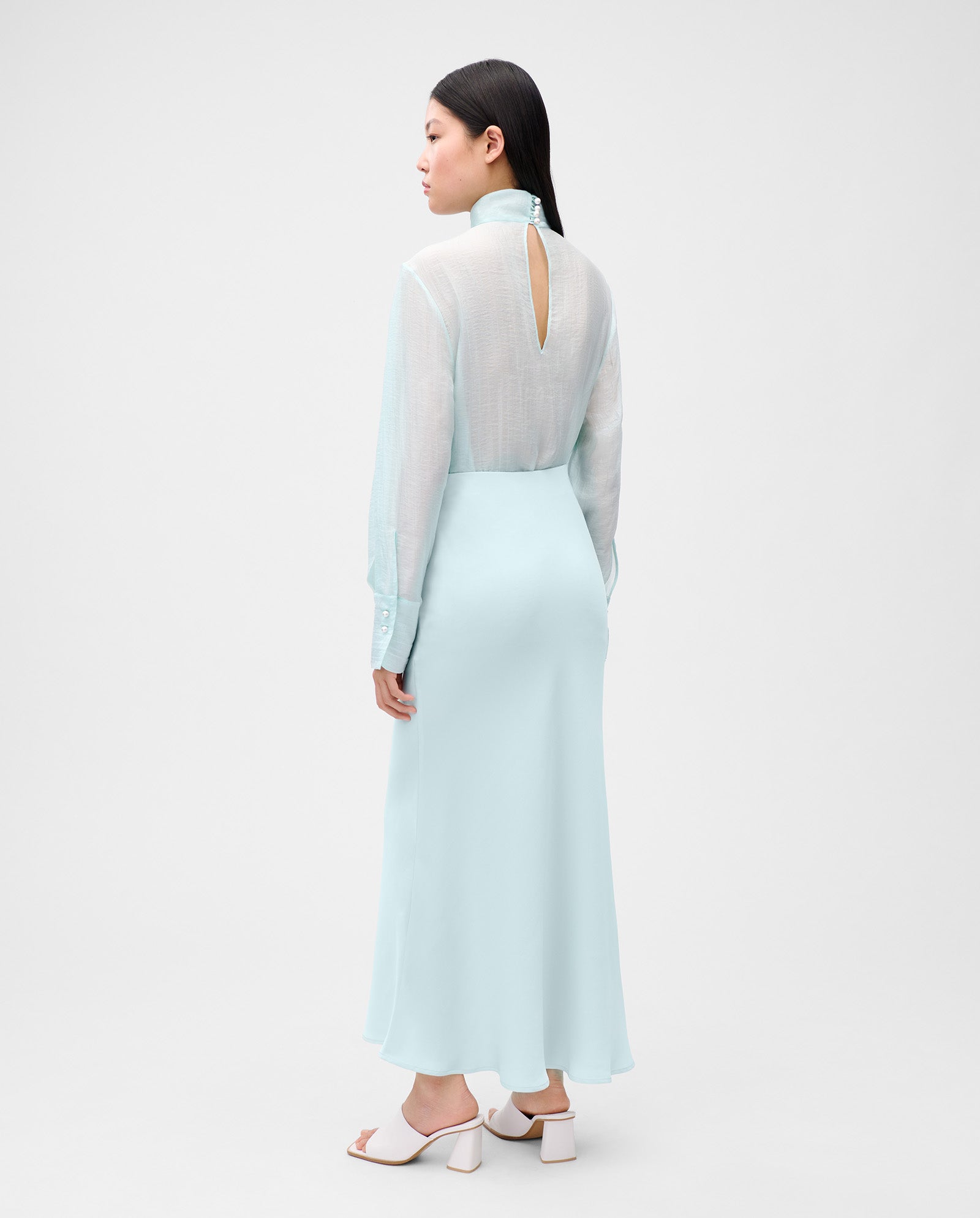 A woman wearing the IVY OAK SUMMER Skirt in light blue satin with long sleeves and ankle length, paired with white heels, stands with her back to the camera.