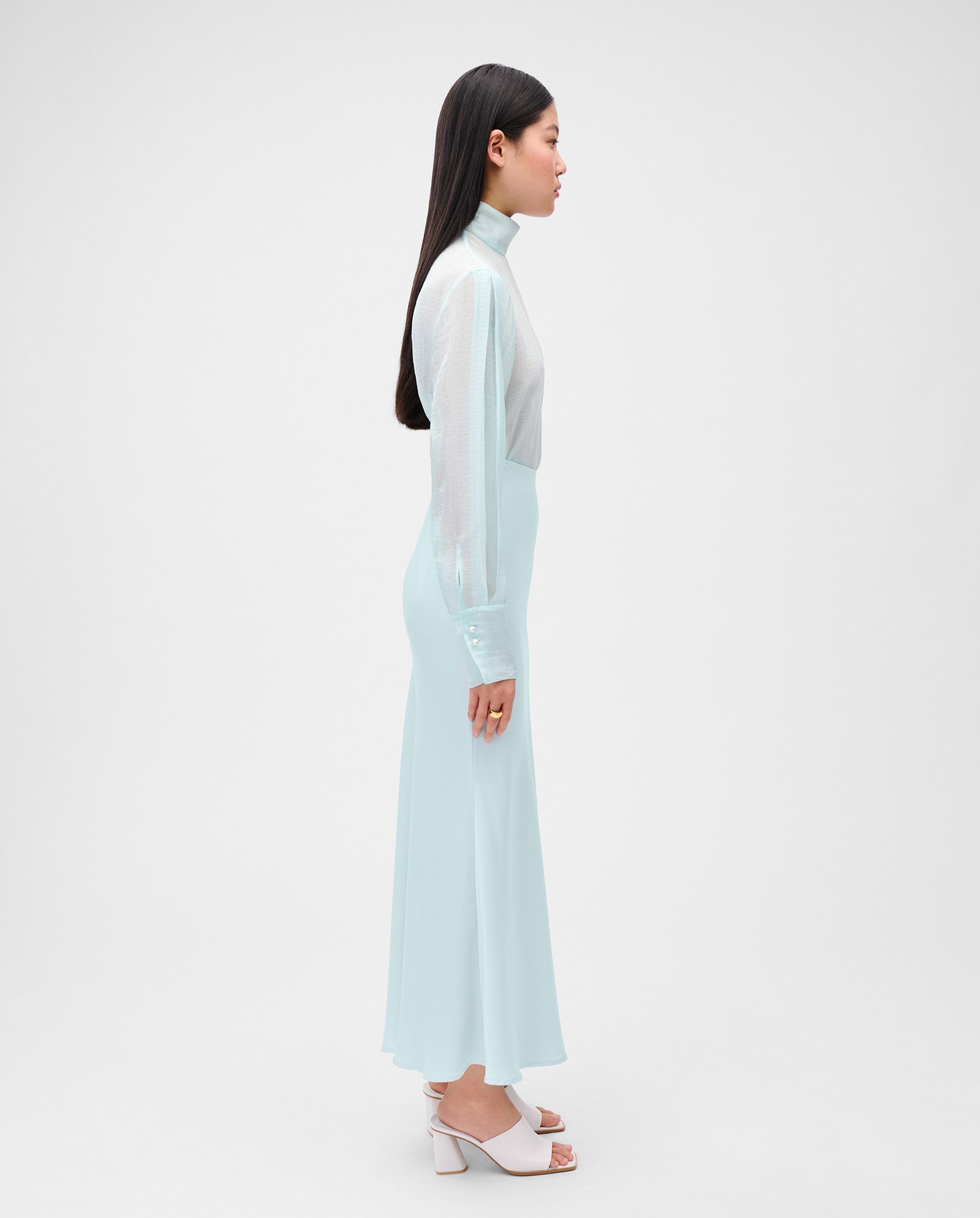Woman in a light blue, ankle-length SUMMER Skirt by IVY OAK, made from satin fabric, paired with a sheer blouse and white heels, standing in profile against a white background.