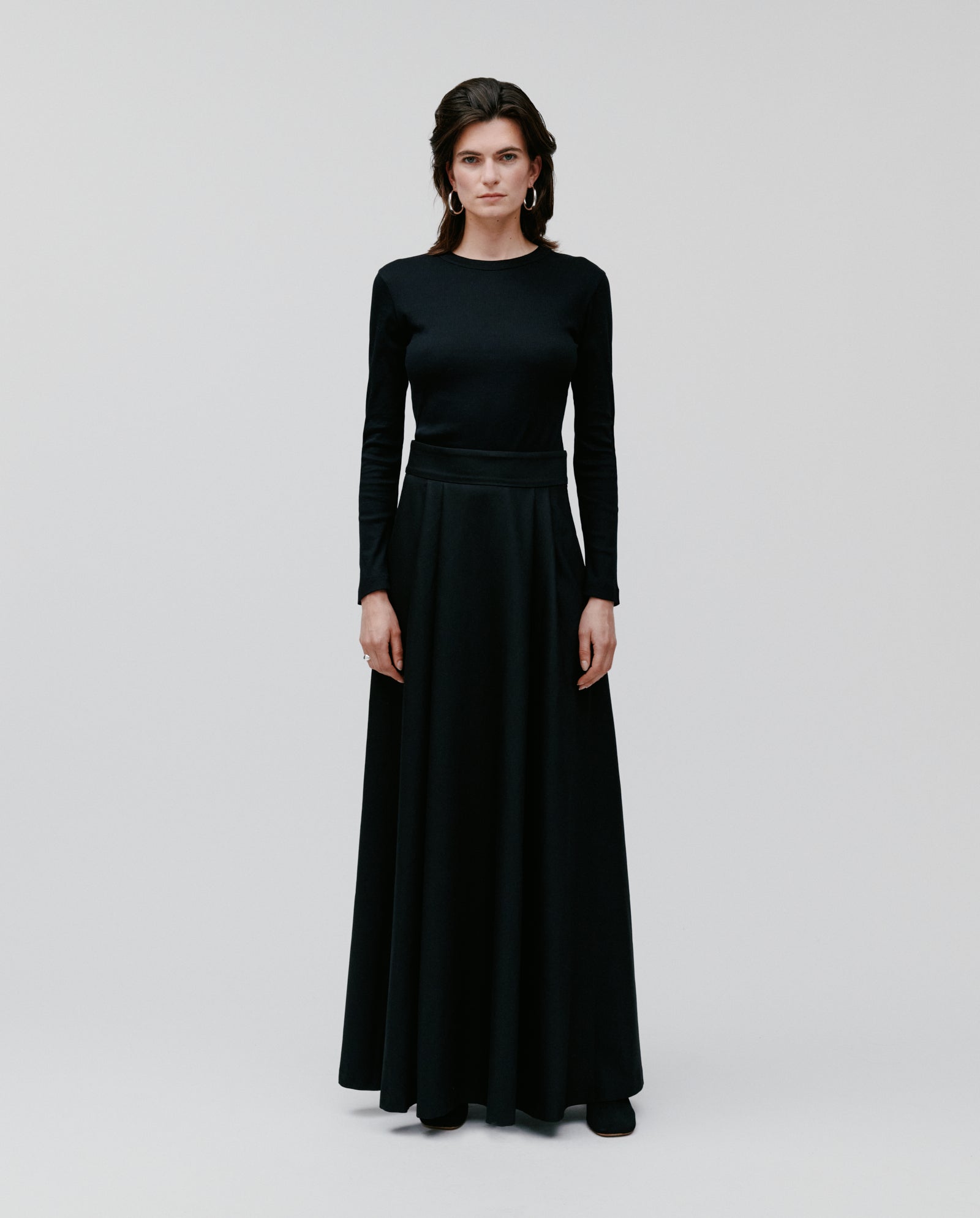 A woman stands against a plain background wearing the IVY OAK SUSANNA Skirt—a floor-length black skirt with graceful pleats and elegant long sleeves.