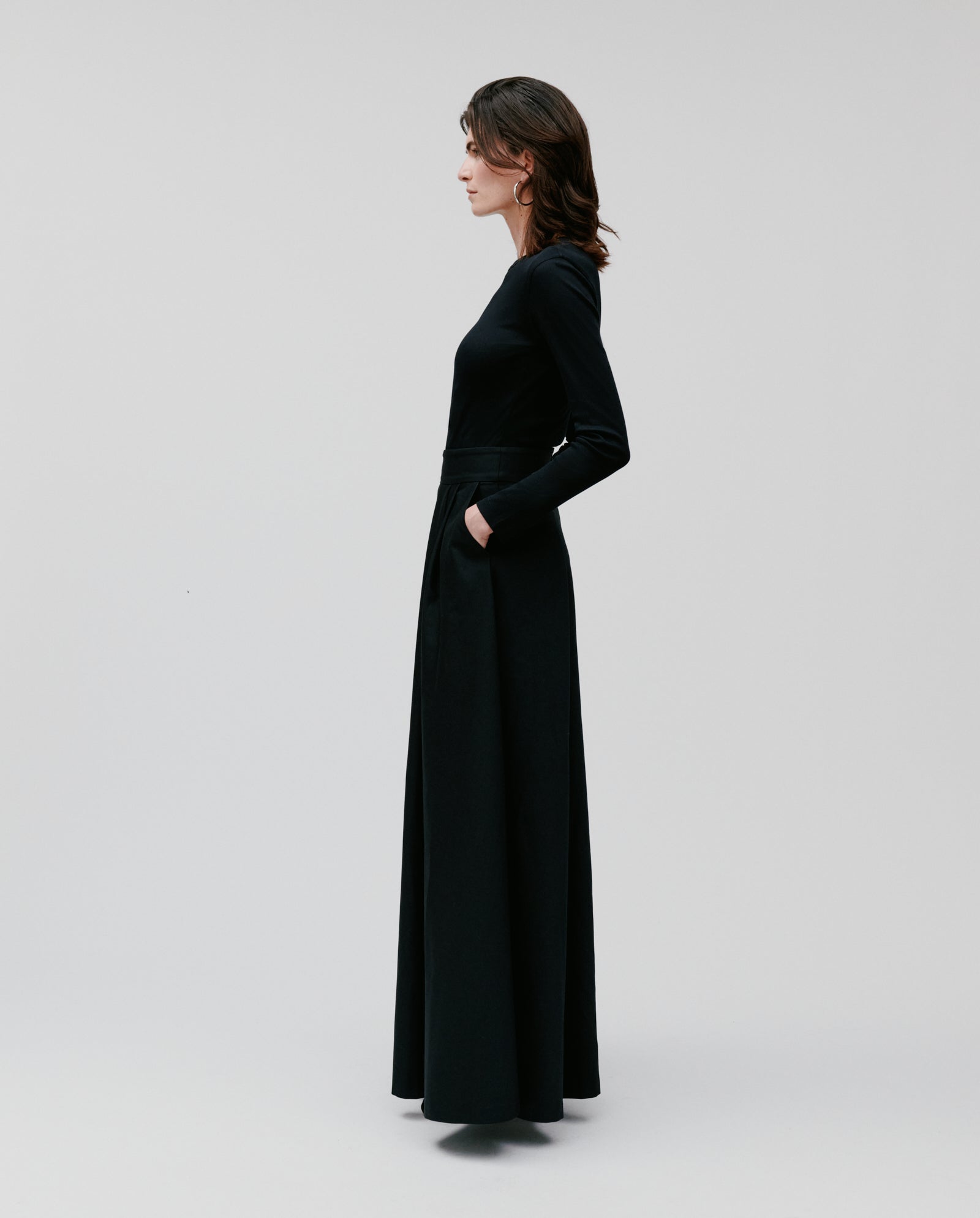 Woman in profile against a plain light background wearing the IVY OAK SUSANNA Skirt, a long black pleated piece paired with a matching long-sleeved top.