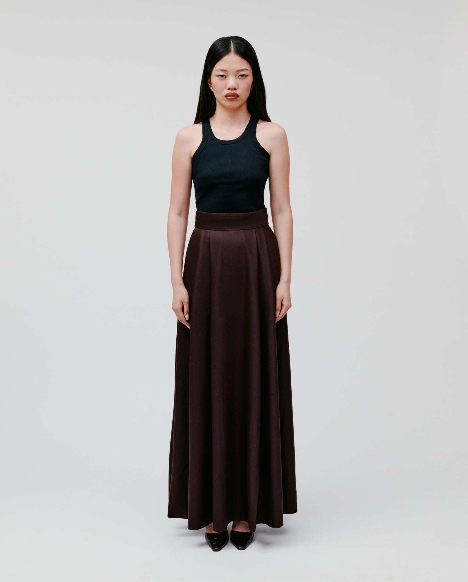 A woman stands against a plain background, wearing a black sleeveless top and the IVY OAK SUSANNA Skirt—a dark brown pleated skirt with pockets.
