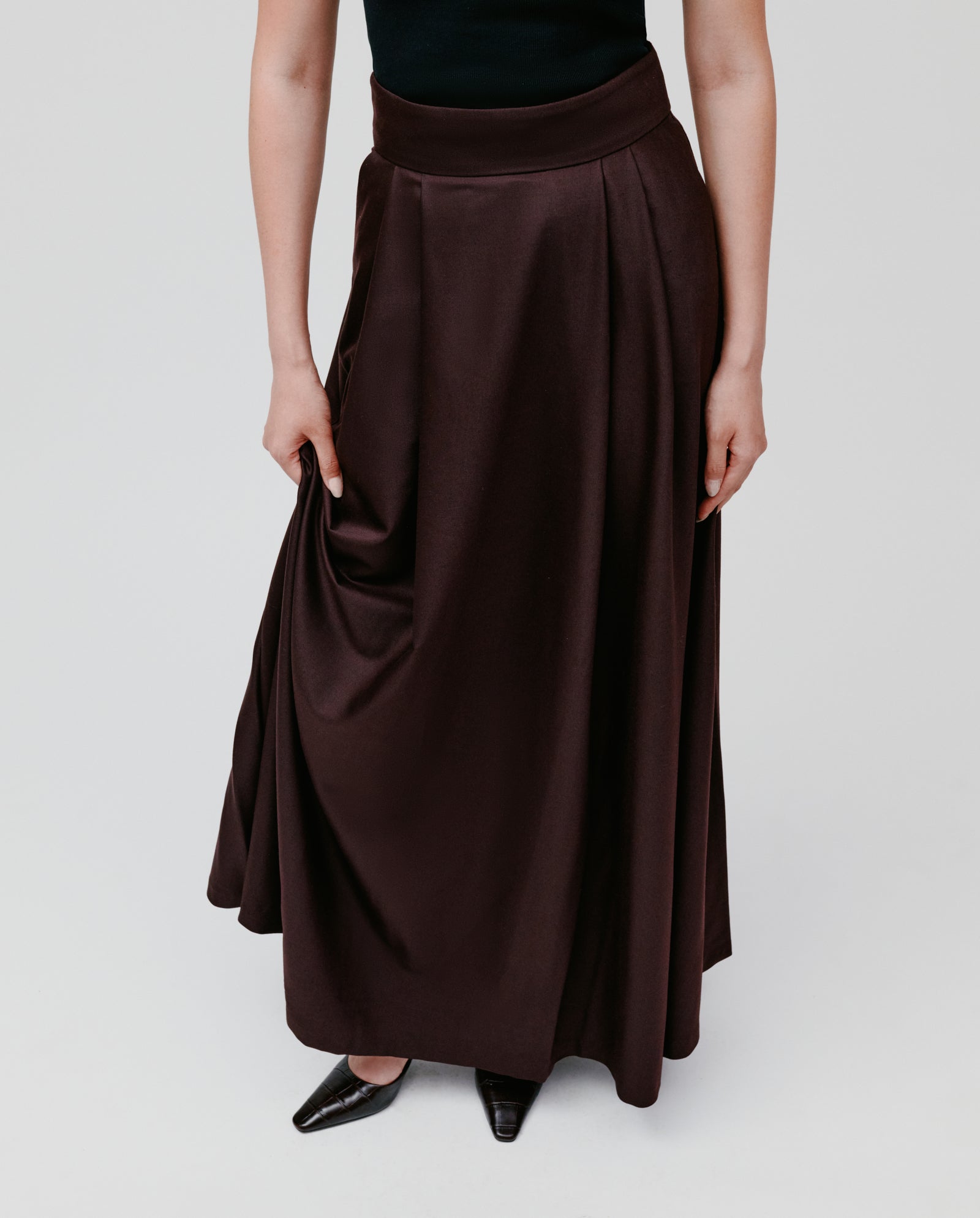 A person wearing a black sleeveless top, the IVY OAK SUSANNA Skirt in dark brown maxi length, and black pointed shoes stands on a white background.