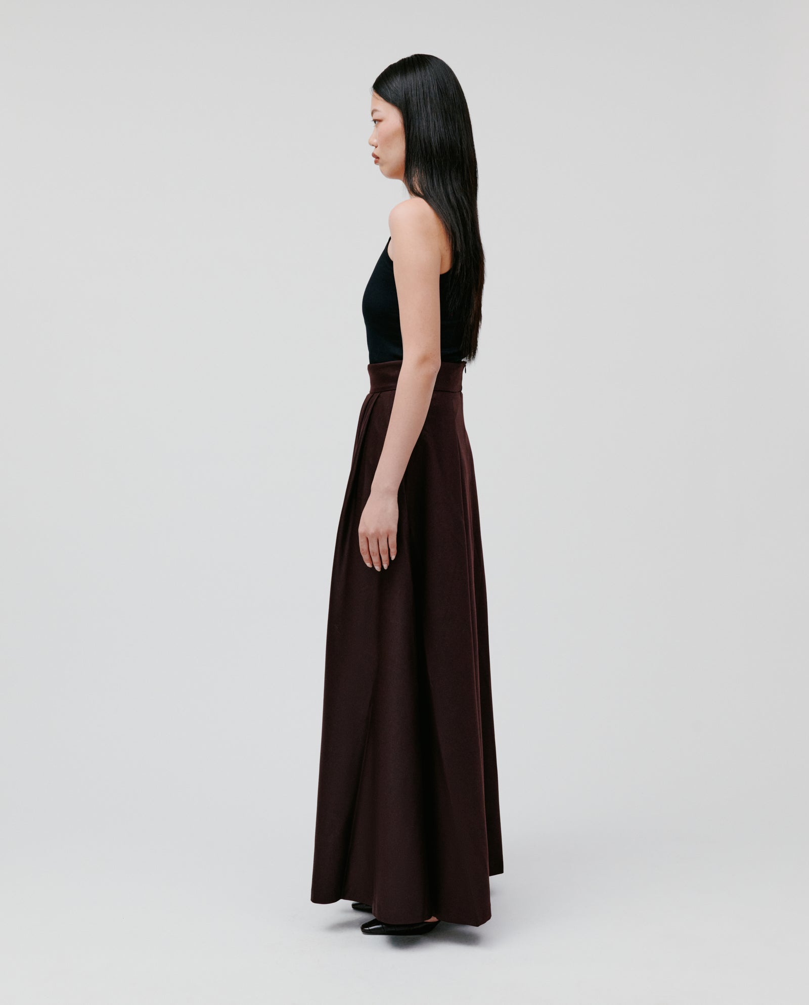 Woman in a black top and the IVY OAK SUSANNA Skirt, a pleated maxi-length piece, stands in profile against a plain light background.