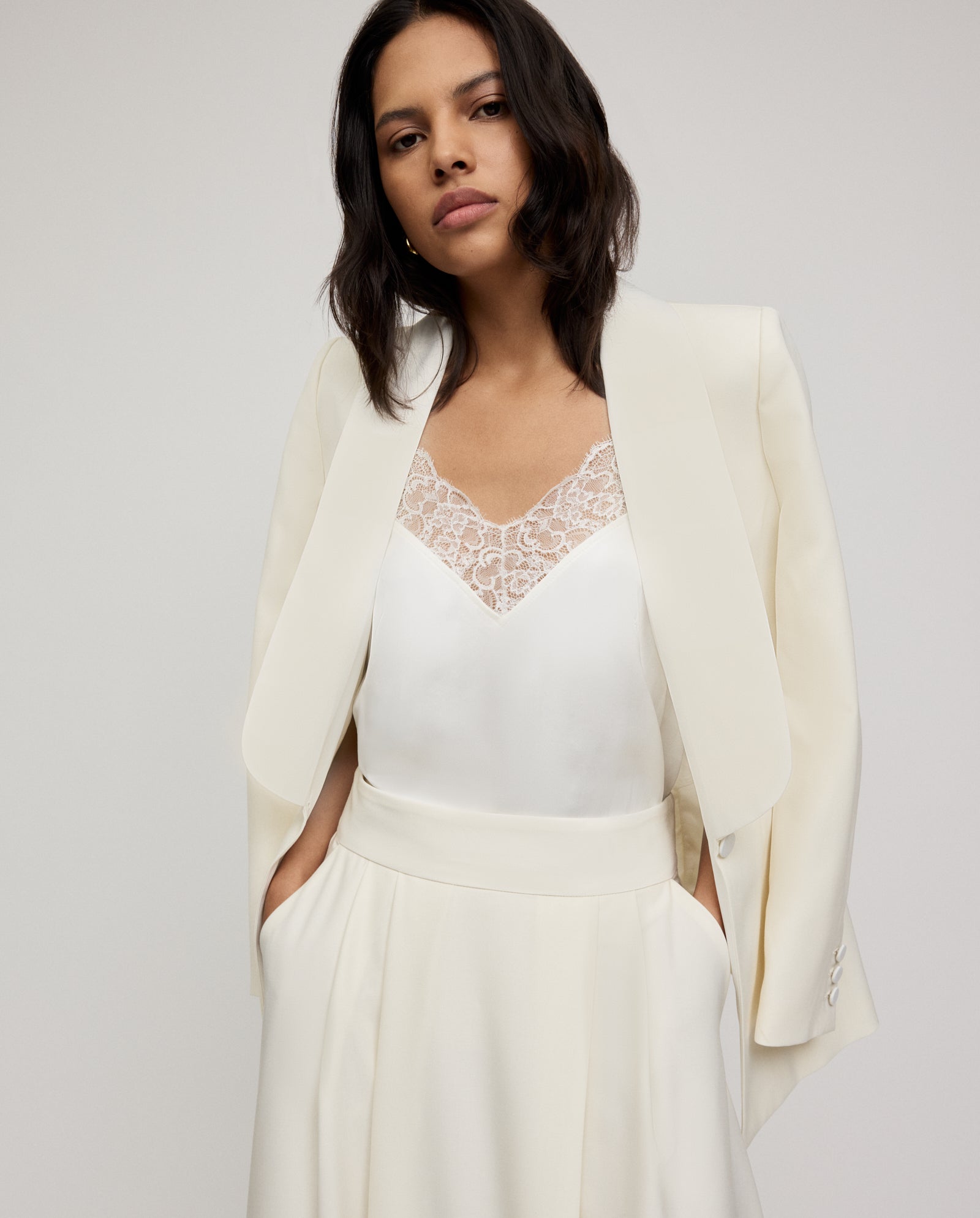 A woman wears the IVY OAK SUSANNA Skirt with an ivory blazer and a lace-trimmed camisole, hands in pockets.