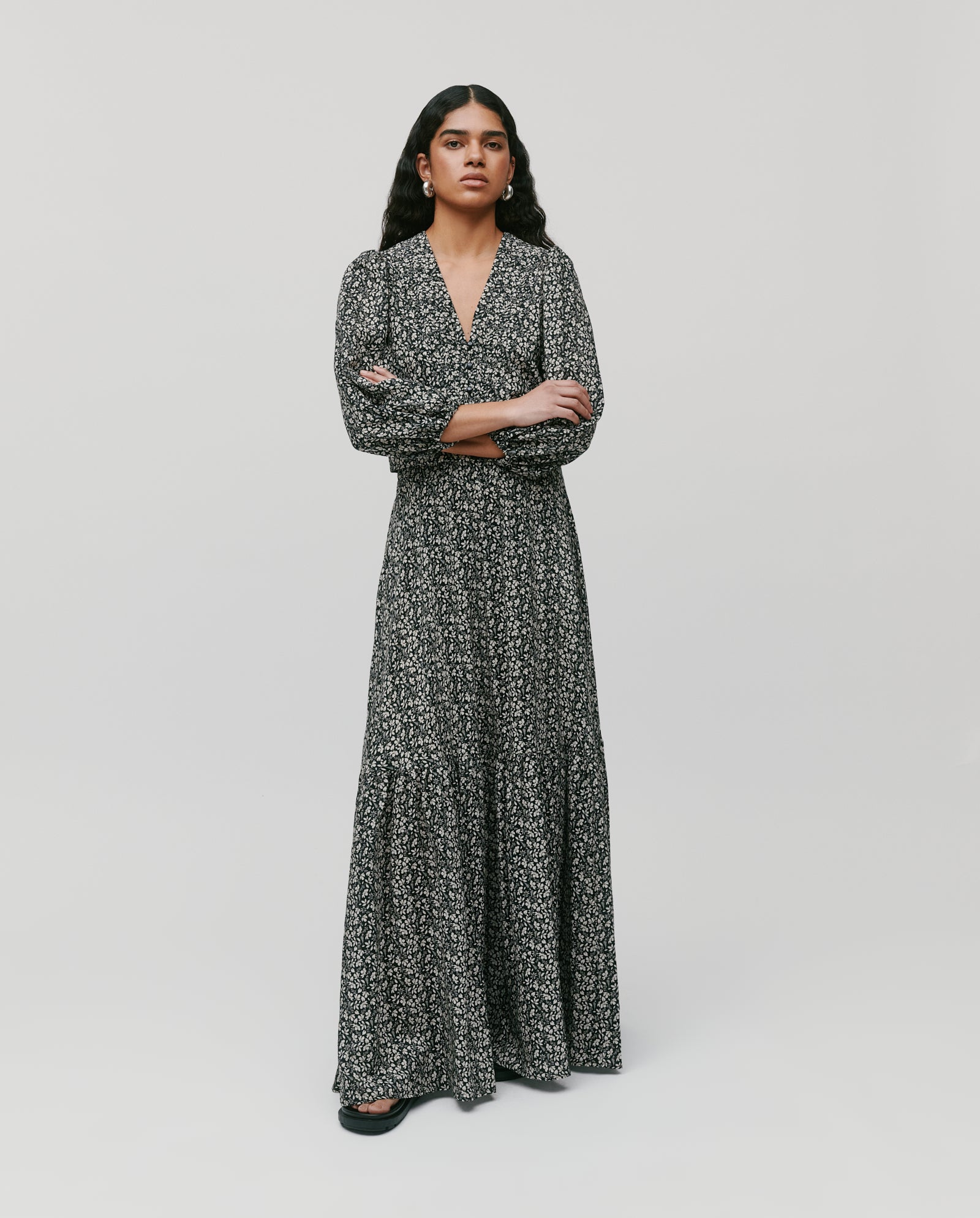 A woman stands with arms crossed against a plain background, wearing the IVY OAK SANJA Skirt—a black and white floral maxi-length design.