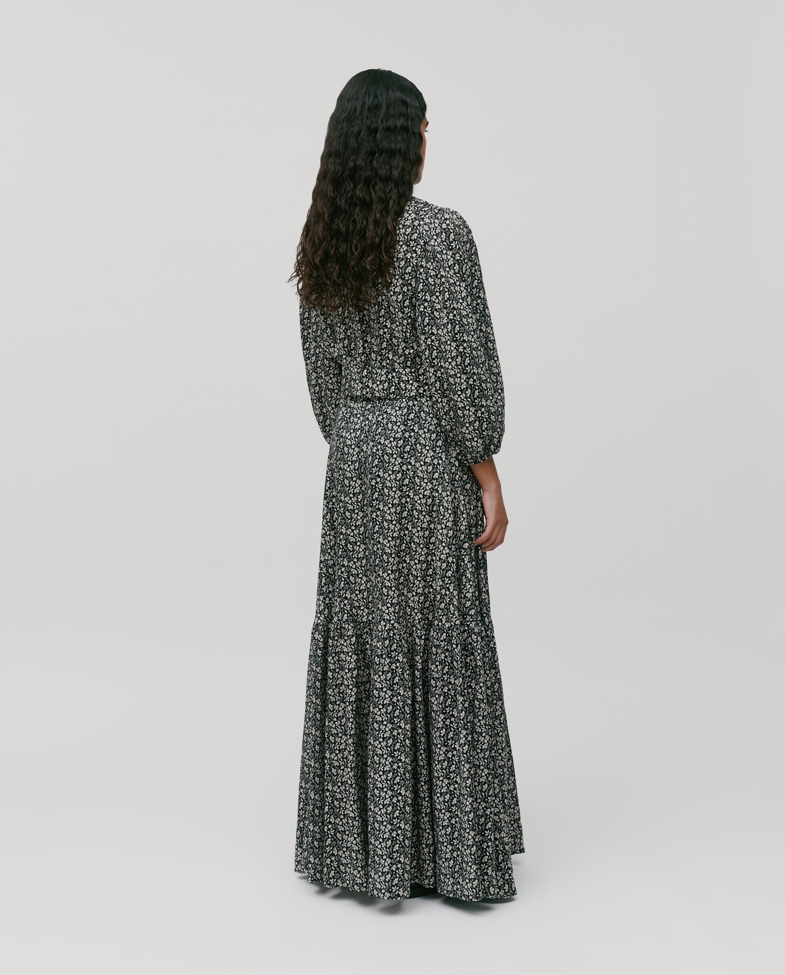 Person with long curly hair seen from behind, wearing the IVY OAK SANJA Skirt—a long black skirt with a white floral pattern and an elastic waistband.