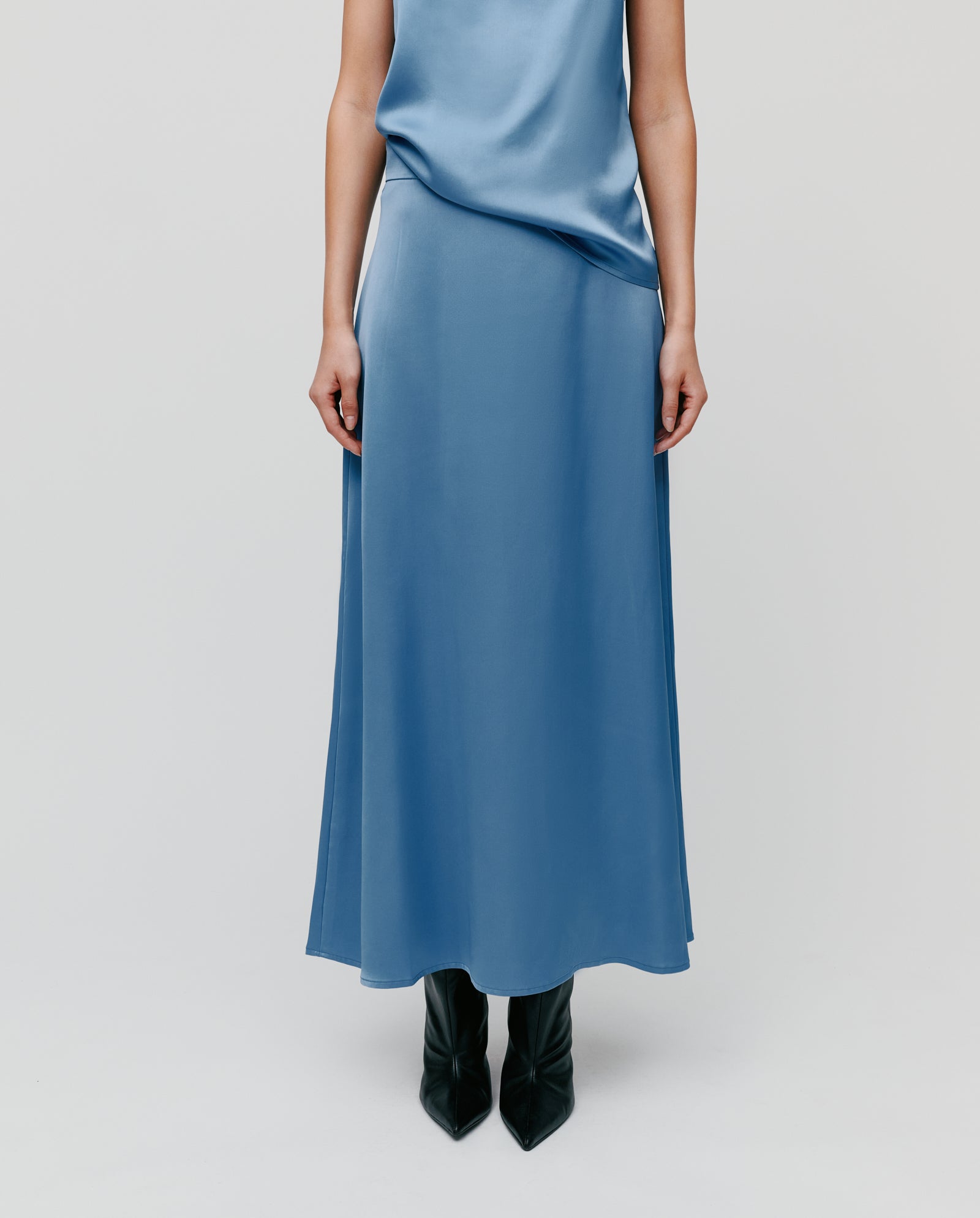 A person wears an ankle-length blue satin IVY OAK SANNA Skirt made with NAIA acetate and black boots, arms relaxed at their sides, shown from the shoulders down.