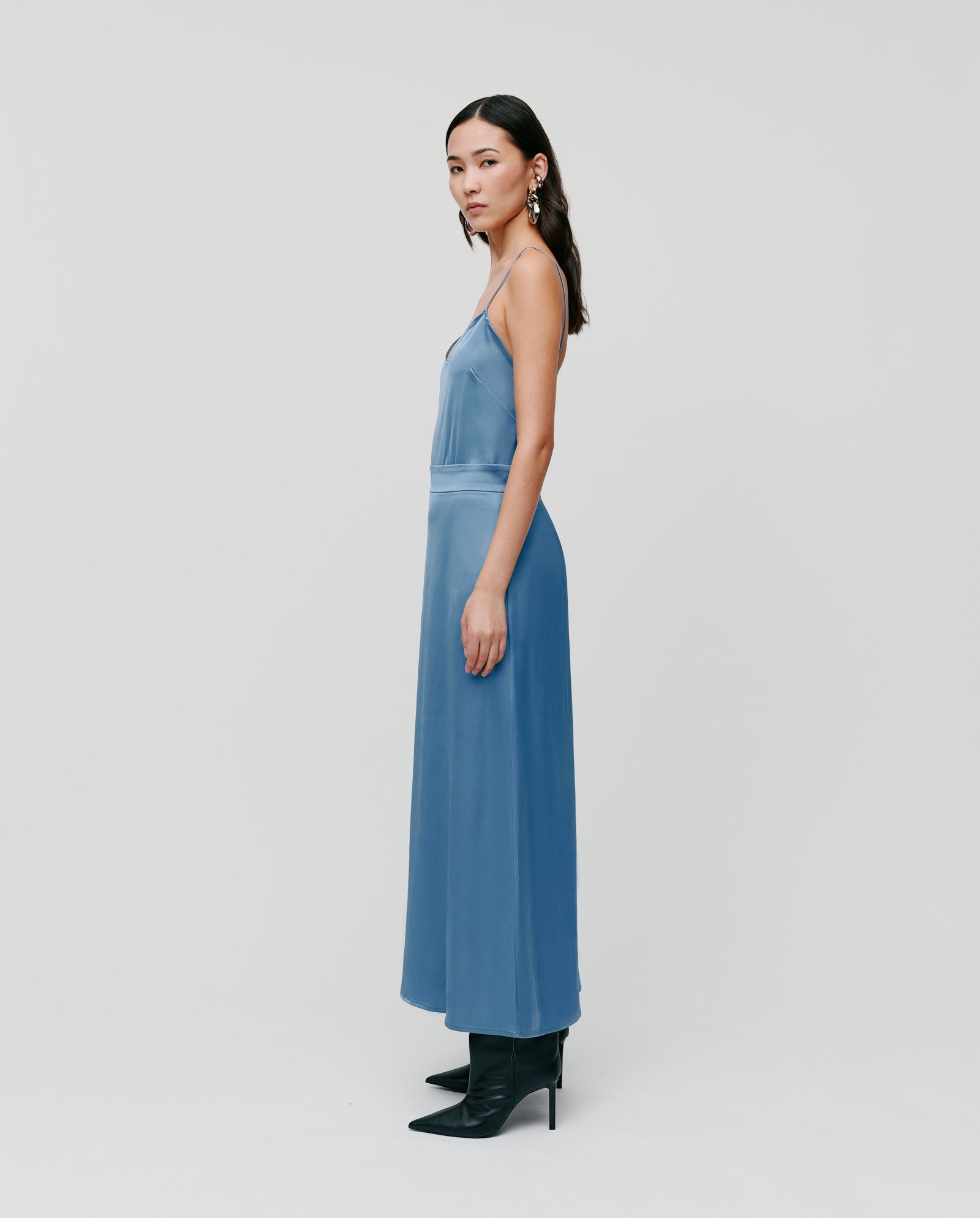 A woman in a blue sleeveless dress styles the IVY OAK SANNA Skirt, ankle-length, with black heeled boots, standing sideways against a plain background.