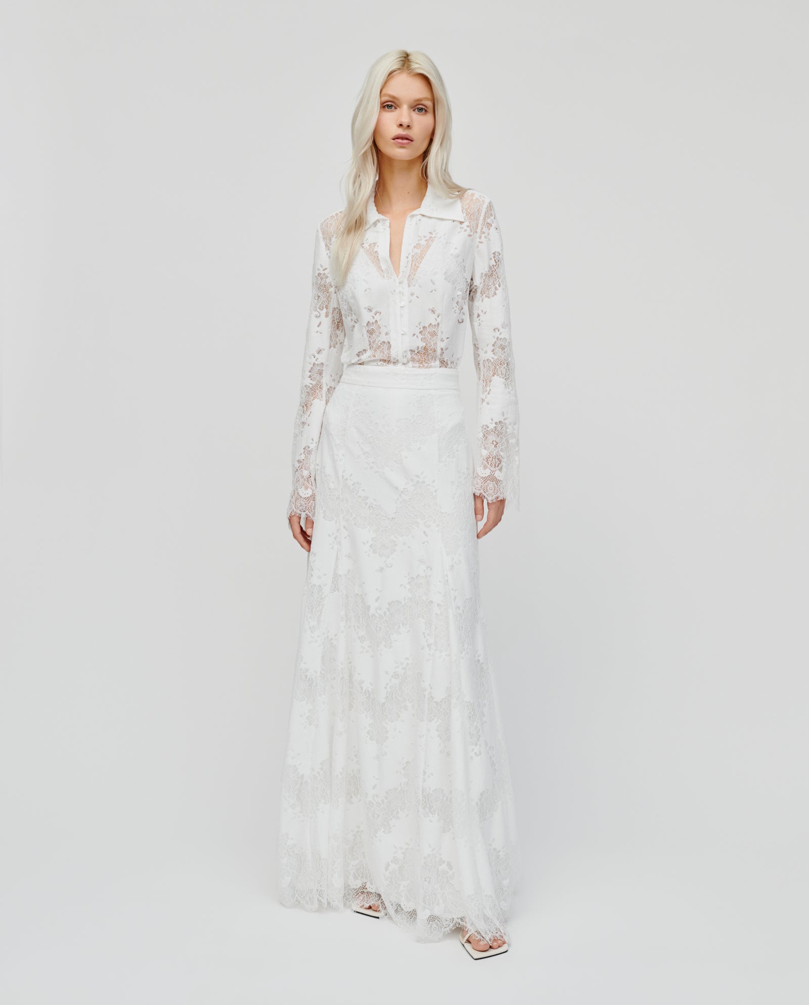 A woman models the IVY OAK SHAYA Lace Skirt, featuring a long, white lace design with a flared maxi silhouette, standing against a plain white background.