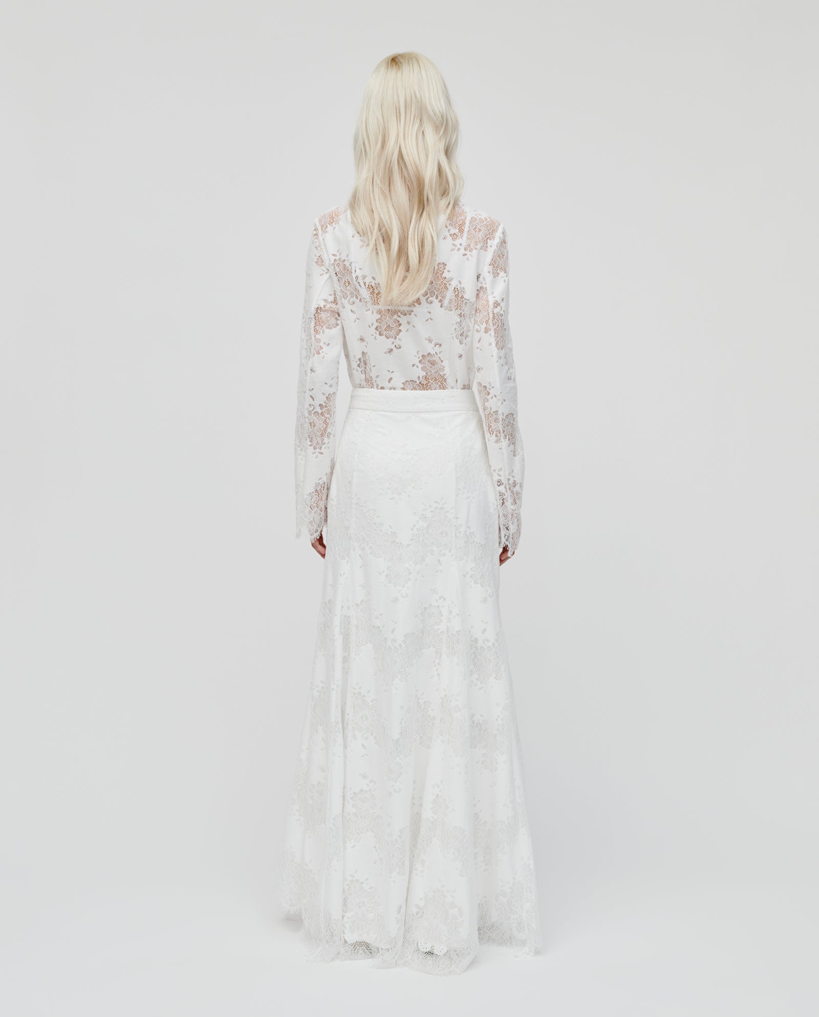 A woman with long blonde hair is shown from the back wearing the IVY OAK SHAYA Lace Skirt, a long-sleeved white lace dress featuring a flared maxi skirt and concealed zipper.