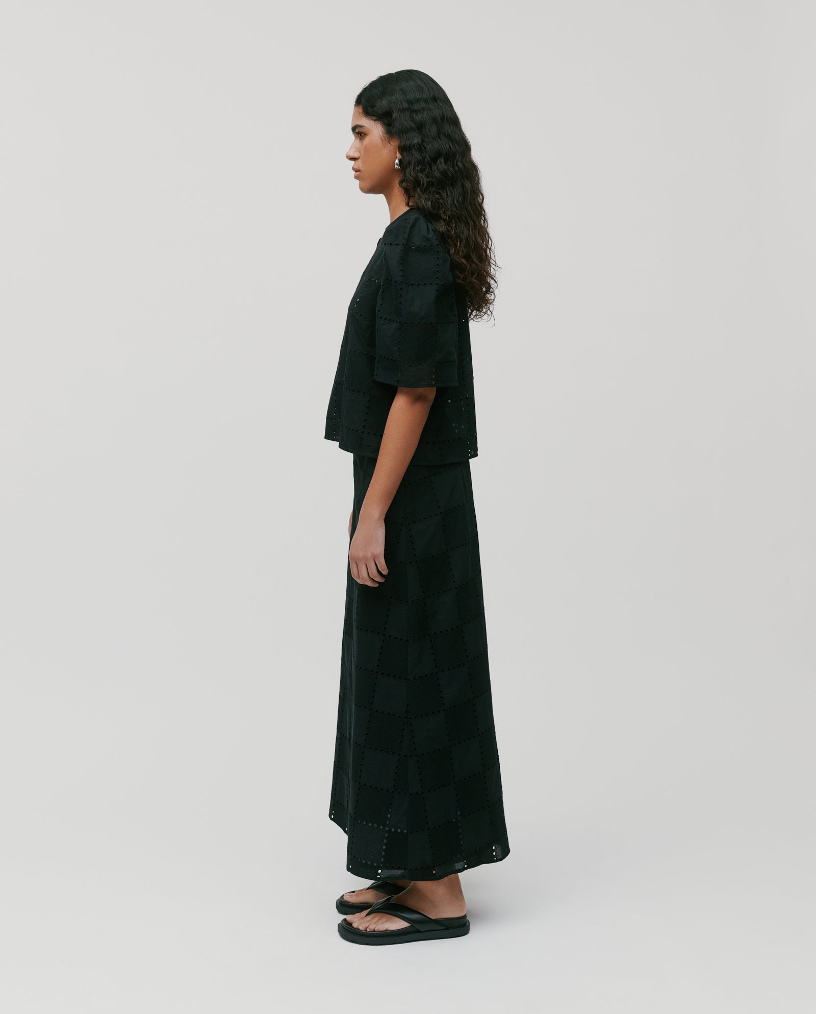 A woman in profile wears a black short-sleeve top, the IVY OAK SAMANTHA Skirt—a broderie anglaise maxi with back zipper closure—and sandals against a plain background.