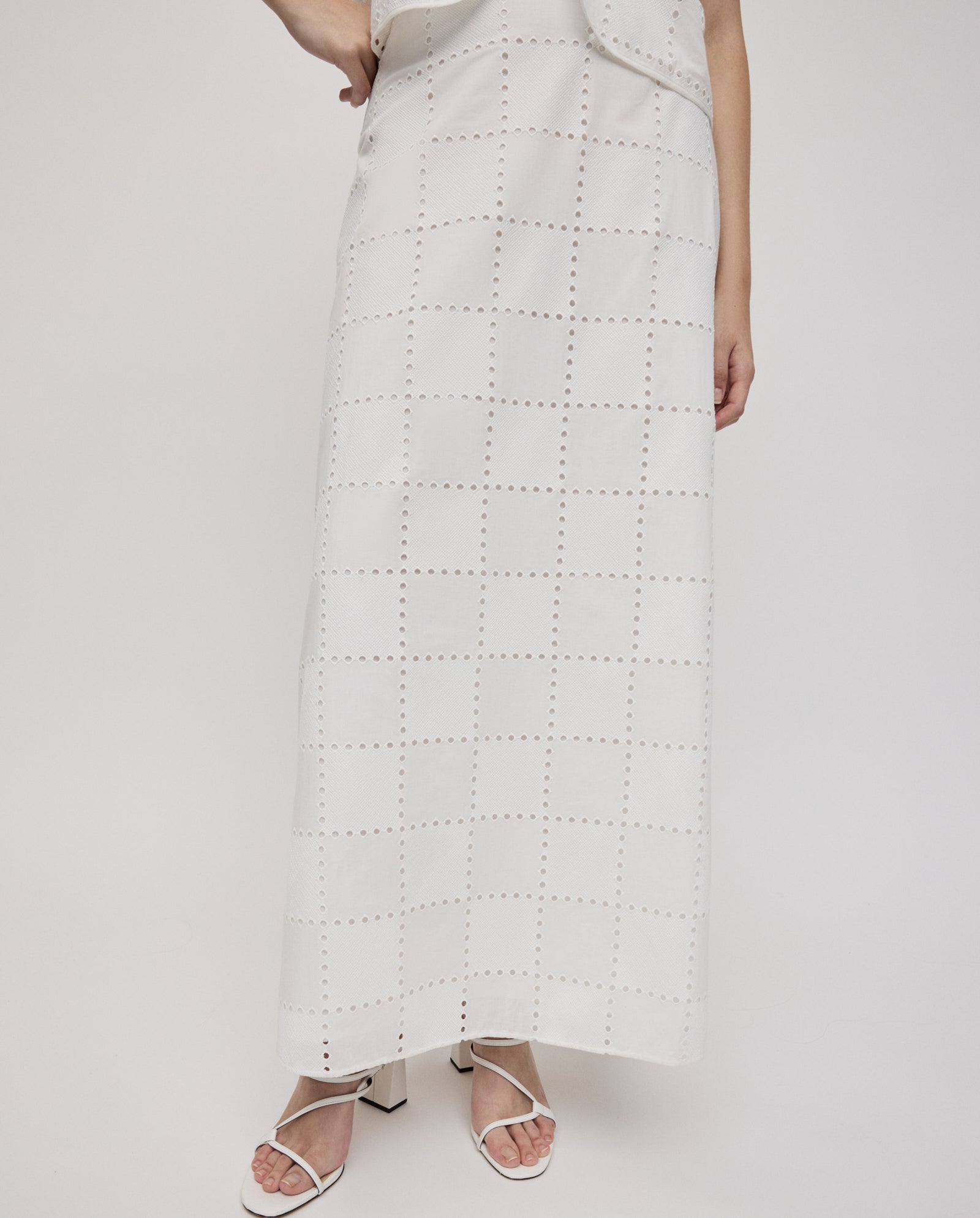 A person wearing the IVY OAK SAMANTHA Skirt, a long white broderie anglaise maxi skirt with a grid pattern, paired with white heeled sandals and featuring a partially visible arm.