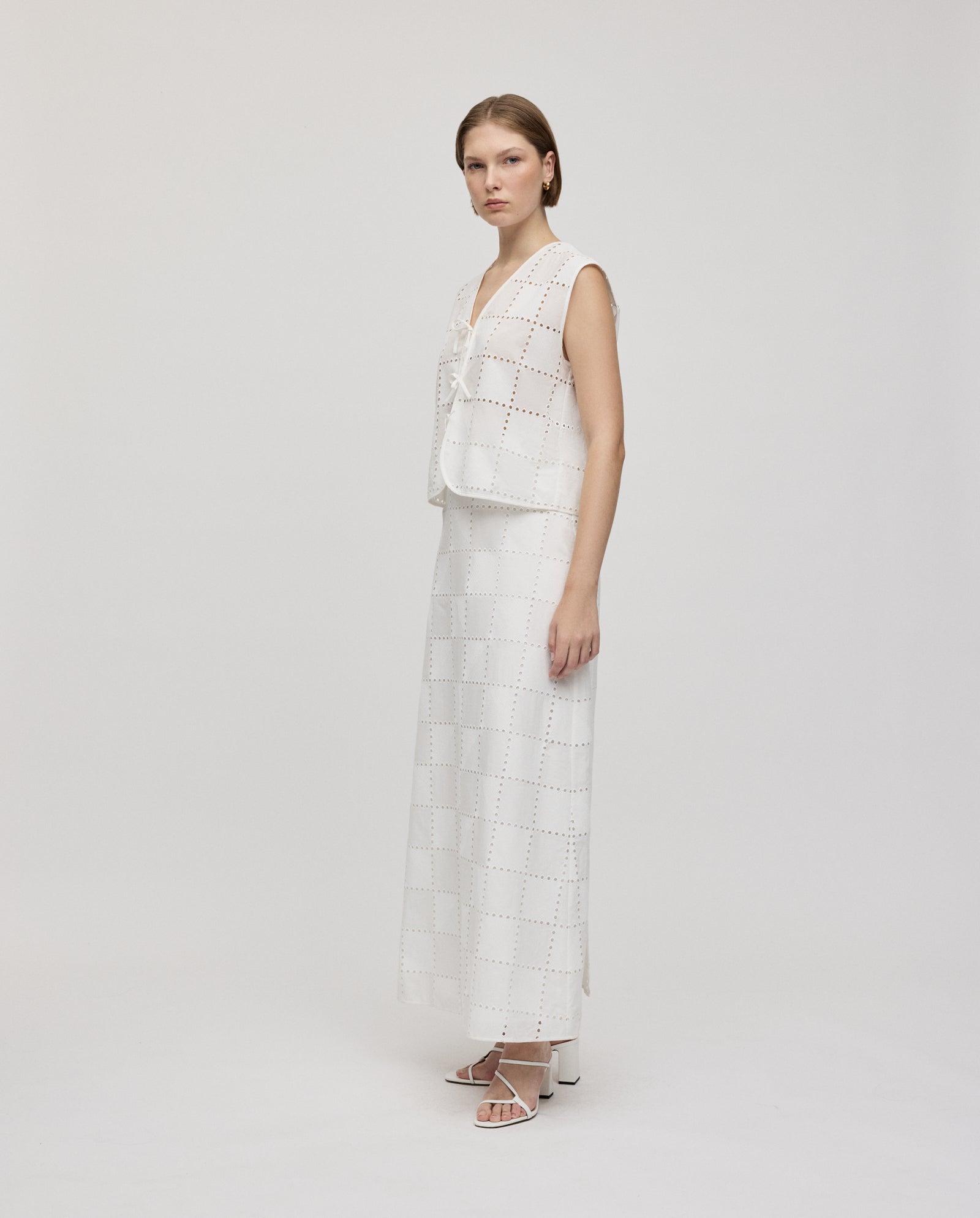A woman wears the IVY OAK SAMANTHA Skirt, a broderie anglaise maxi with square cut-outs, paired with a sleeveless white top, standing on a white background.