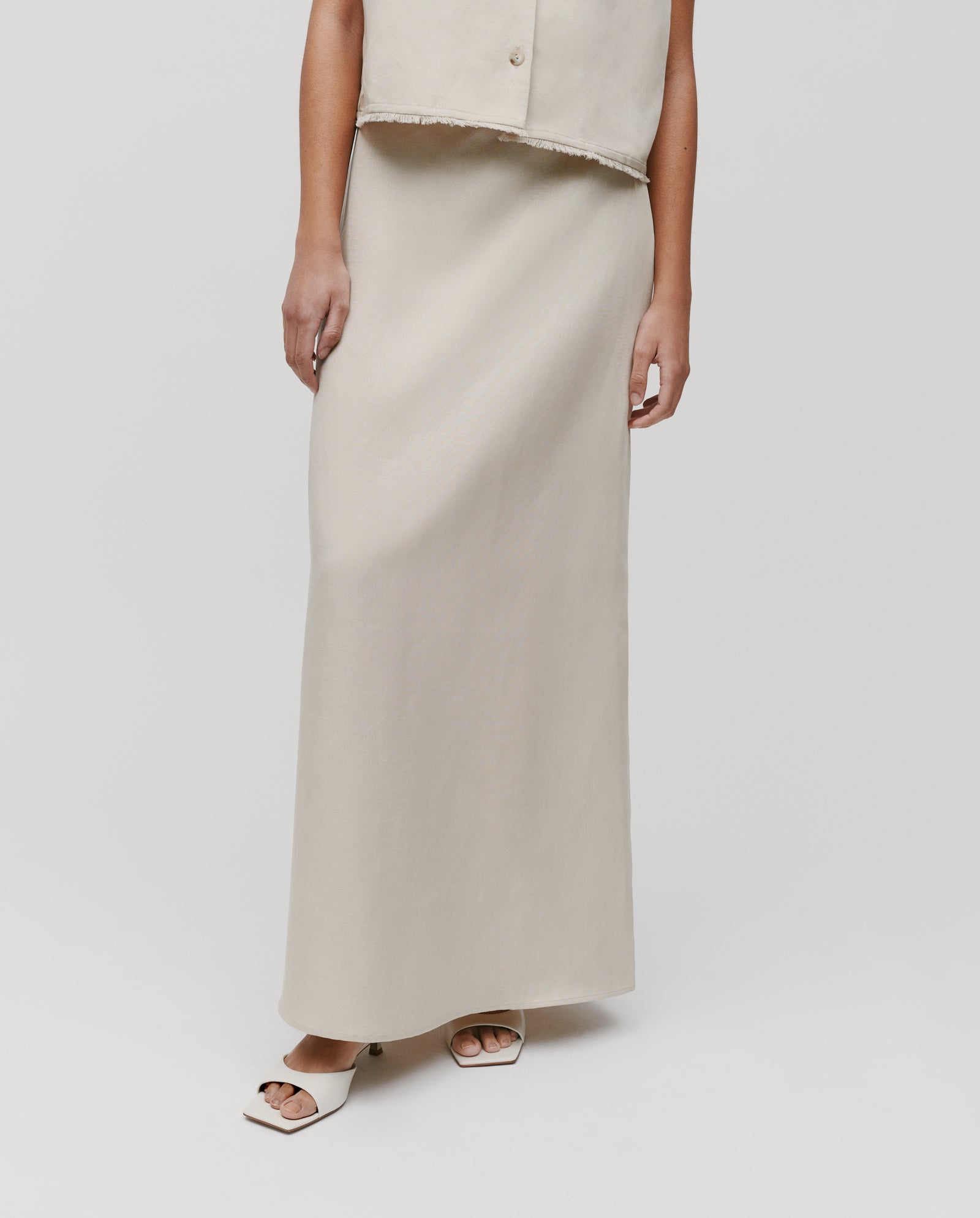 A woman wears the IVY OAK SAMANTHA ANN Skirt in beige with a matching sleeveless top and cream open-toe sandals, set against a white background.