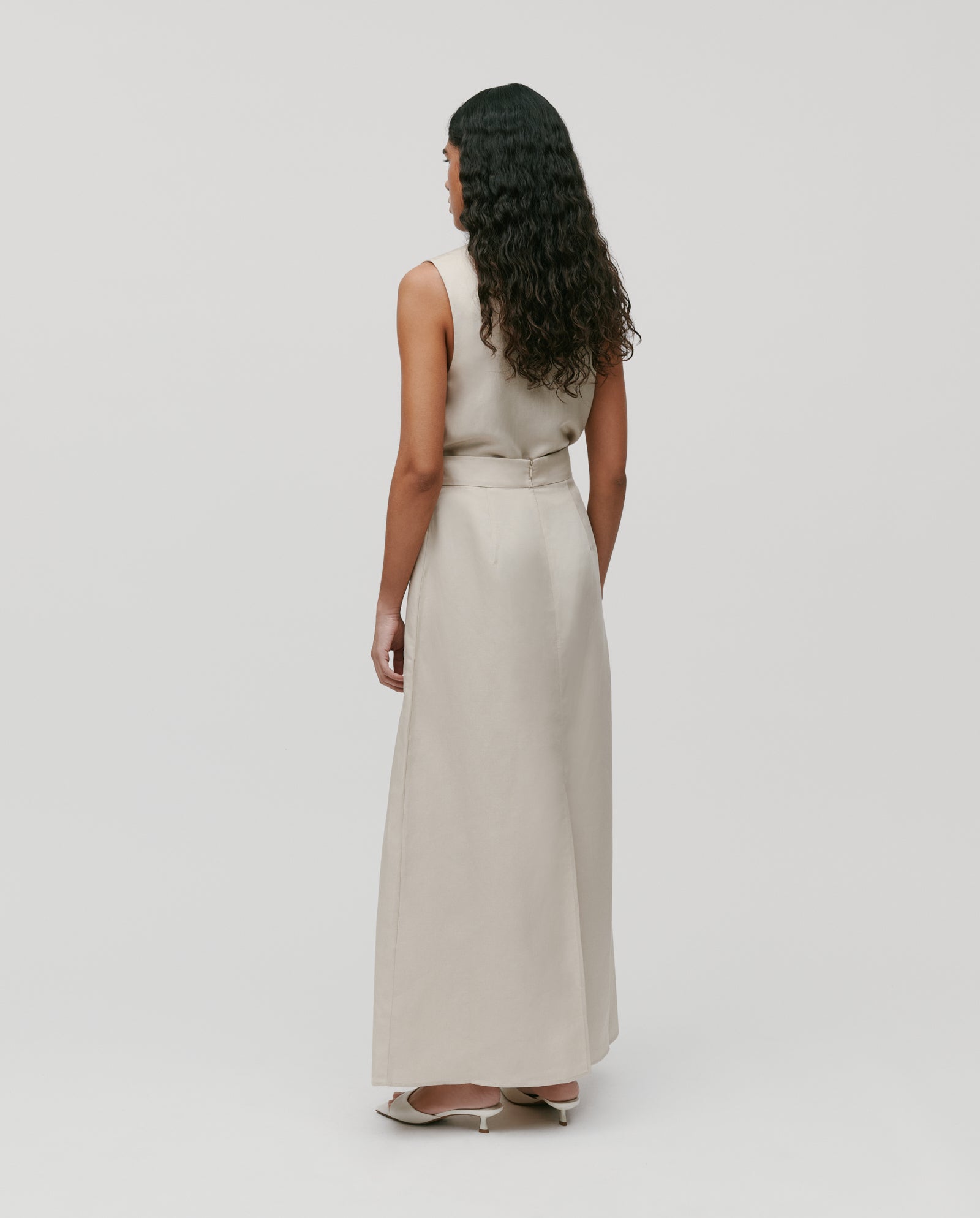 Woman with long curly hair wears a sleeveless beige maxi dress and the IVY OAK SAMANTHA ANN Skirt, paired with heels, standing facing away.