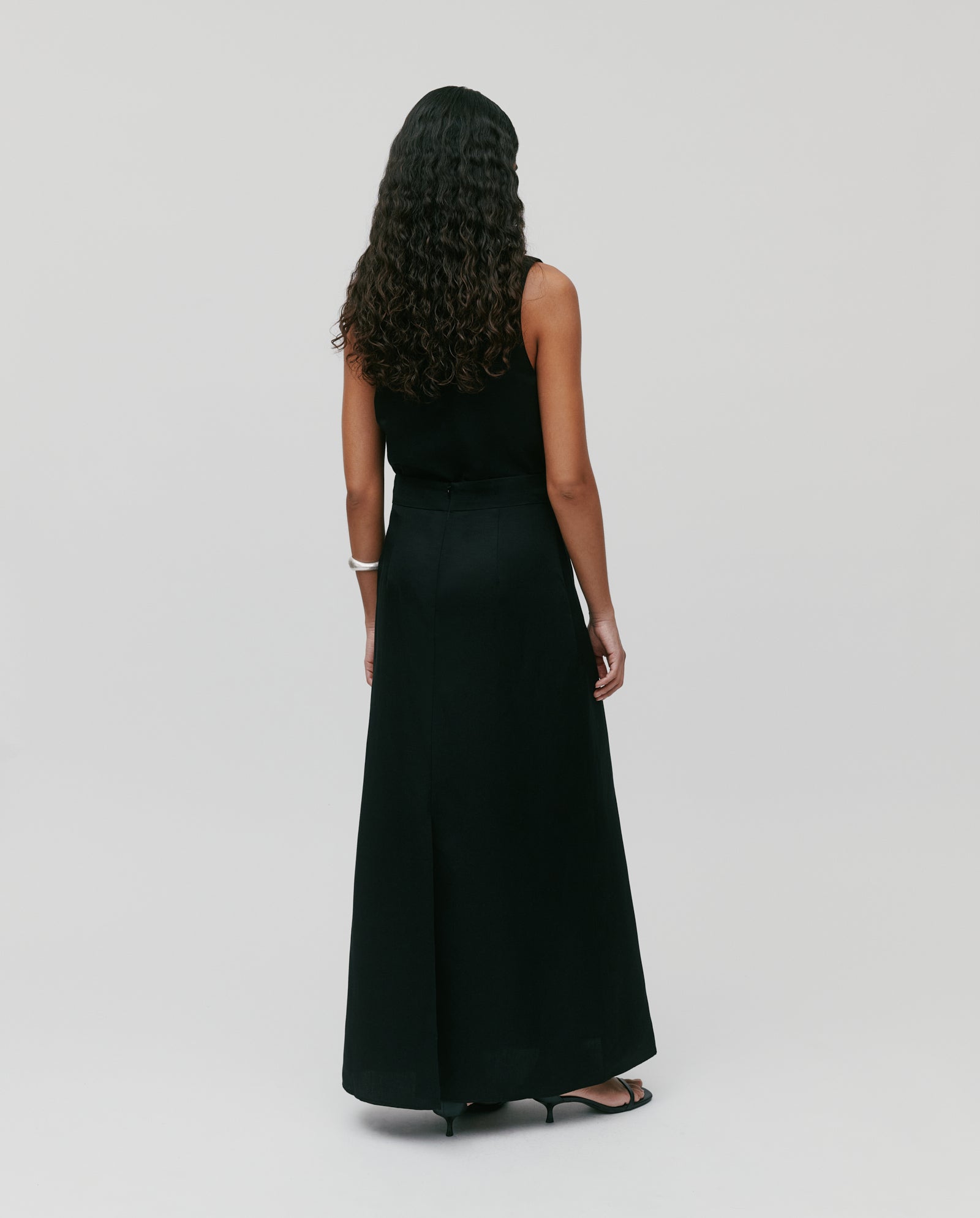 A woman stands with her back to the camera against a plain background, wearing the IVY OAK SAMANTHA ANN Skirt—a long black linen-blend skirt.