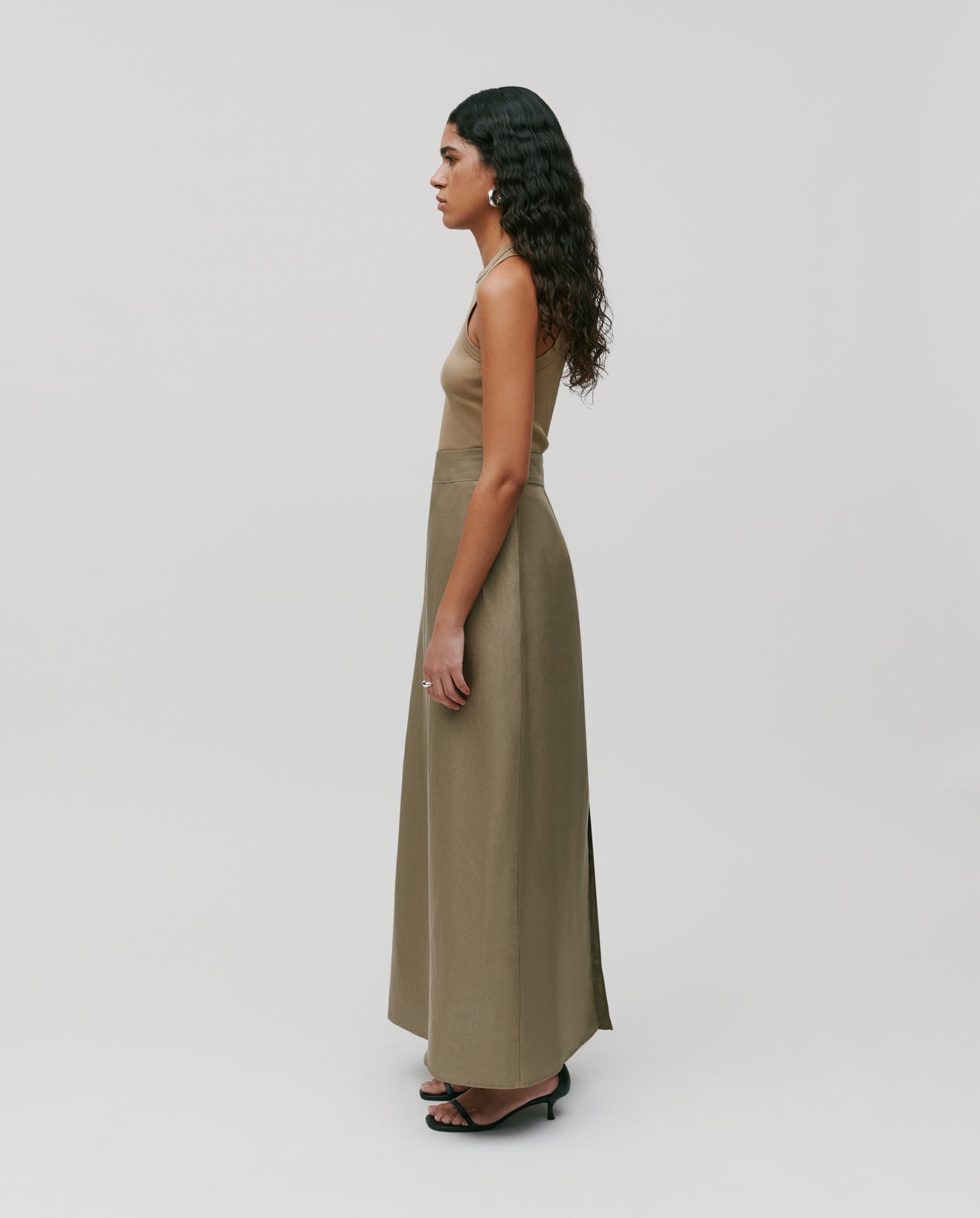 A woman in a sleeveless brown top and an IVY OAK SAMANTHA ANN Skirt, made from a soft linen blend, stands sideways against a plain light background.