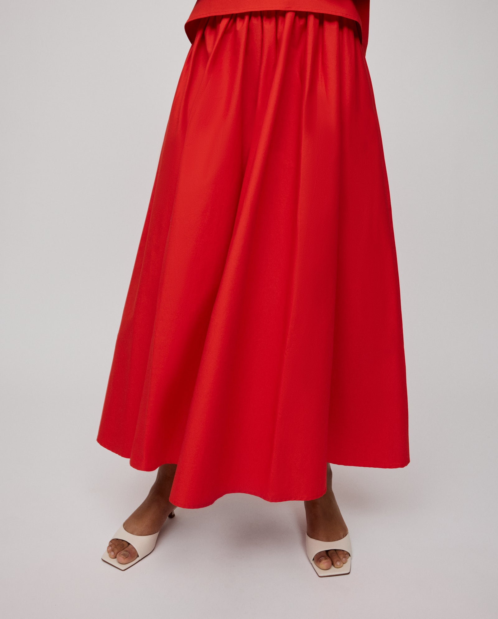 Model wears the IVY OAK SIENNA Skirt, a voluminous, bright red cotton piece with a defined waistband, paired with white open-toe heels against a plain background.