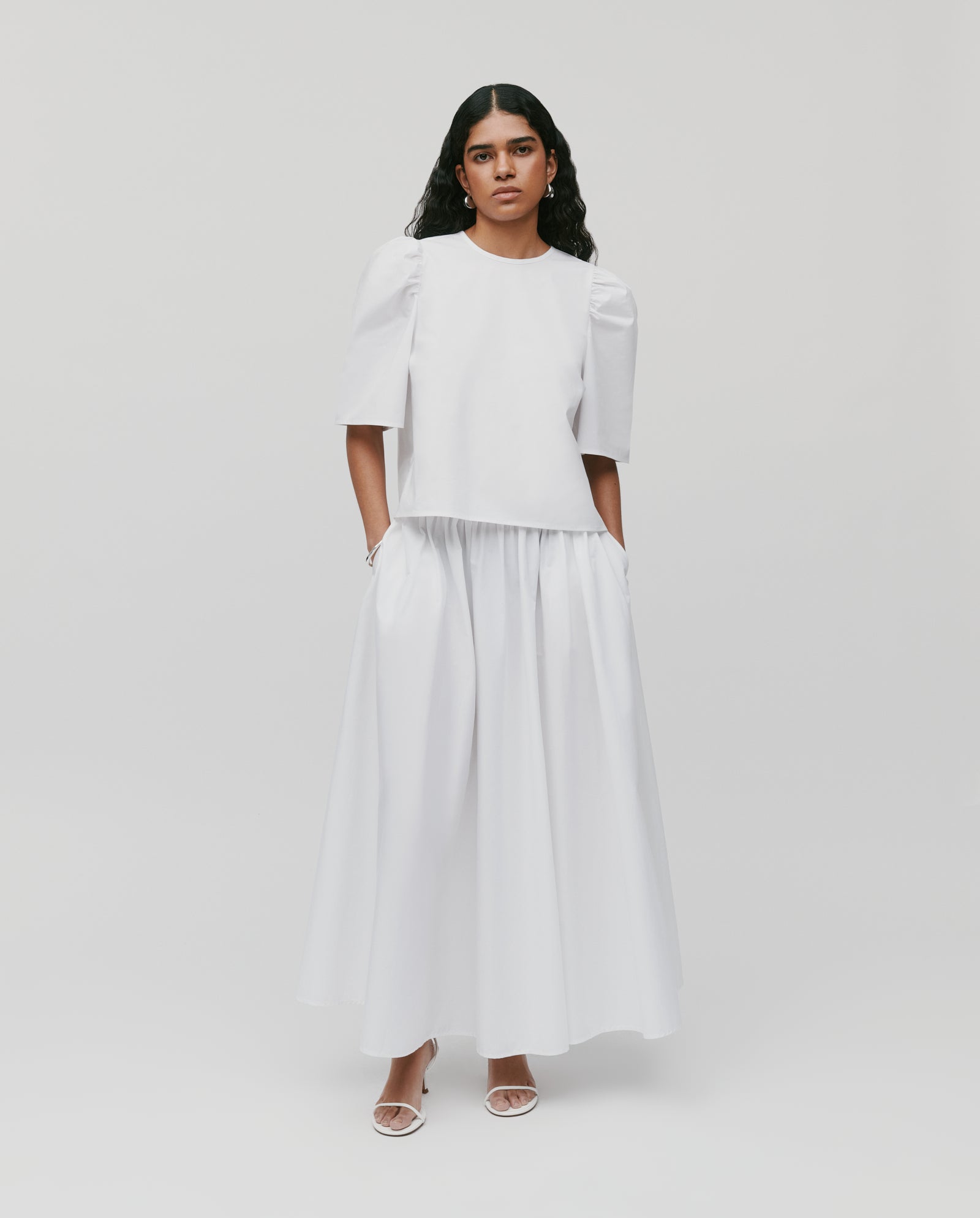 A woman wears the IVY OAK SIENNA Skirt—a voluminous, long cotton skirt with a defined waistband—paired with a white short-sleeve top, standing against a plain light background.