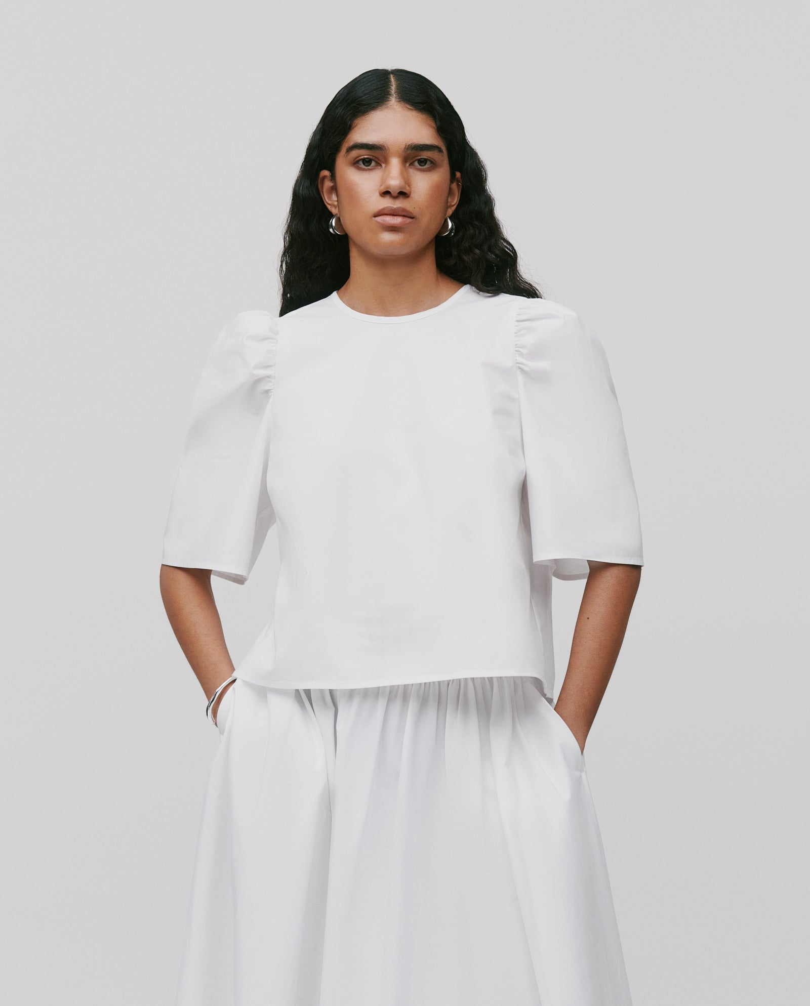 A woman stands with hands in pockets wearing the IVY OAK SIENNA Skirt— a voluminous, long cotton piece with a defined waistband—paired with a white puff-sleeve top against a plain background.