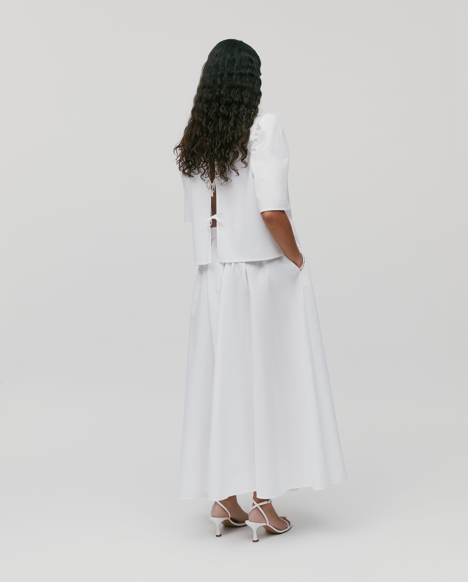 A woman faces away from the camera, wearing a white short-sleeve top paired with the IVY OAK SIENNA Skirt—a voluminous, long cotton style featuring a defined waistband—set against a plain background.