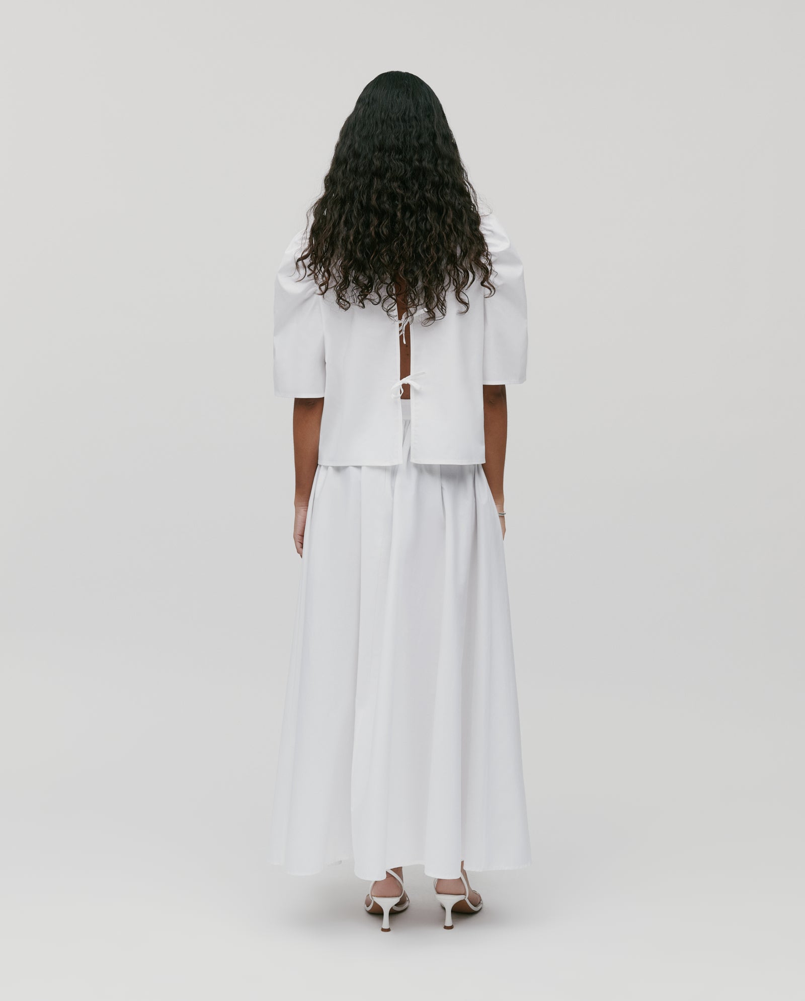 A woman with long curly hair wears the IVY OAK SIENNA Skirt—a voluminous white cotton piece with a defined waistband—as she stands in heels, facing away against a plain background.