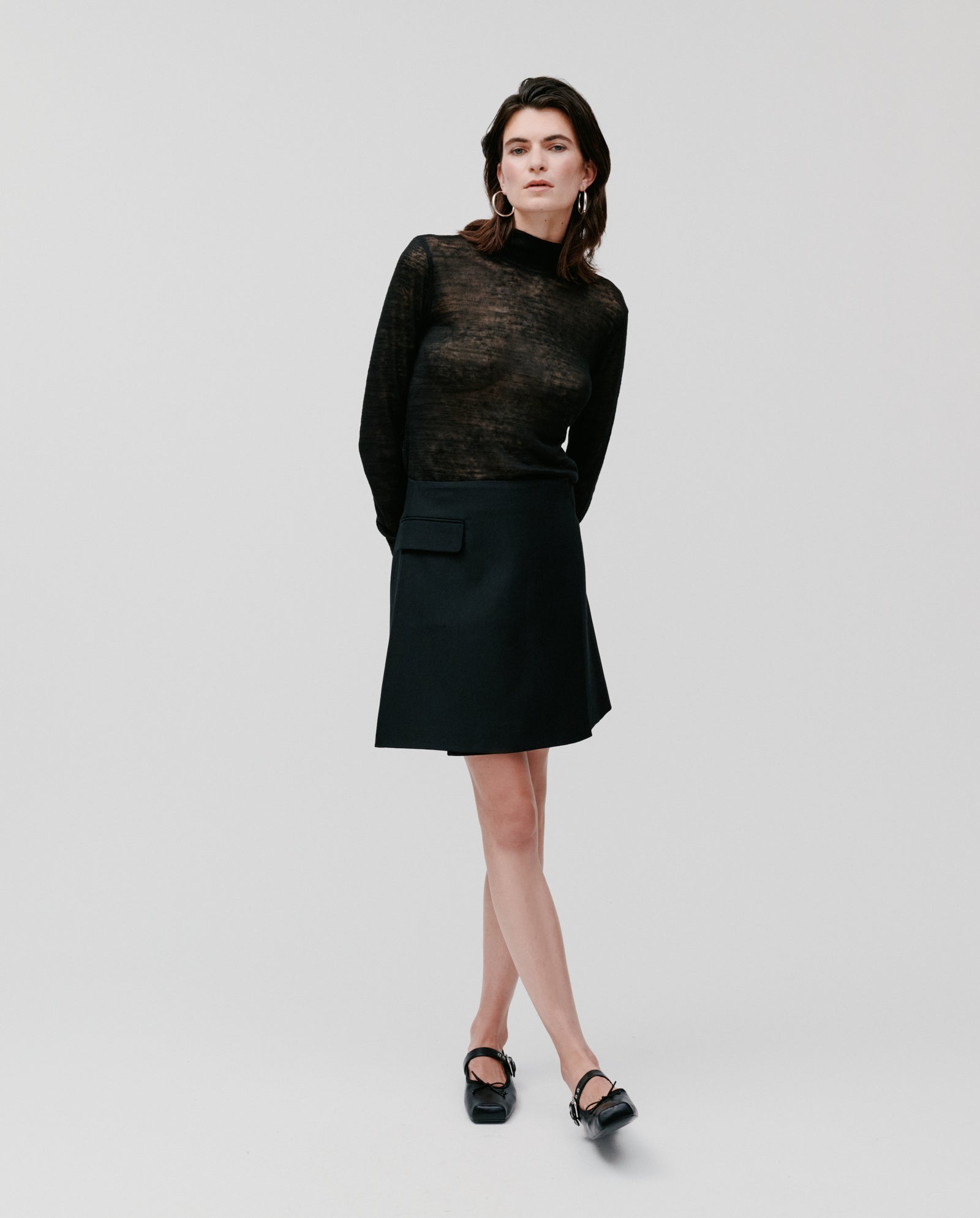 A woman stands with hands behind her back against a plain white background, wearing a black sheer top and the IVY OAK STEVIE Skirt.