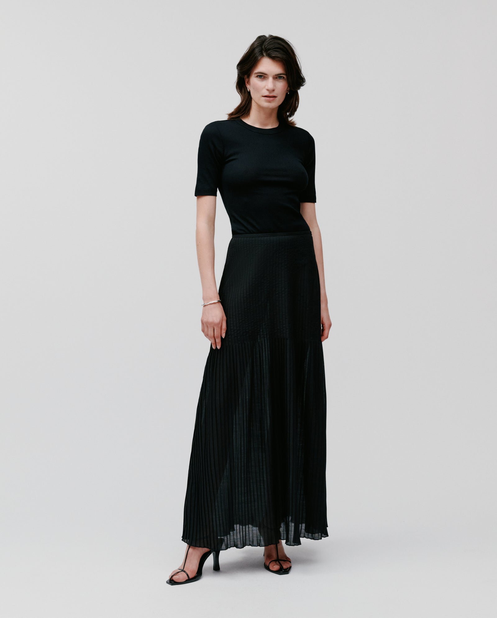 A woman in a black short-sleeve top and the IVY OAK SALENA Skirt, paired with black heeled sandals, stands against a plain background.
