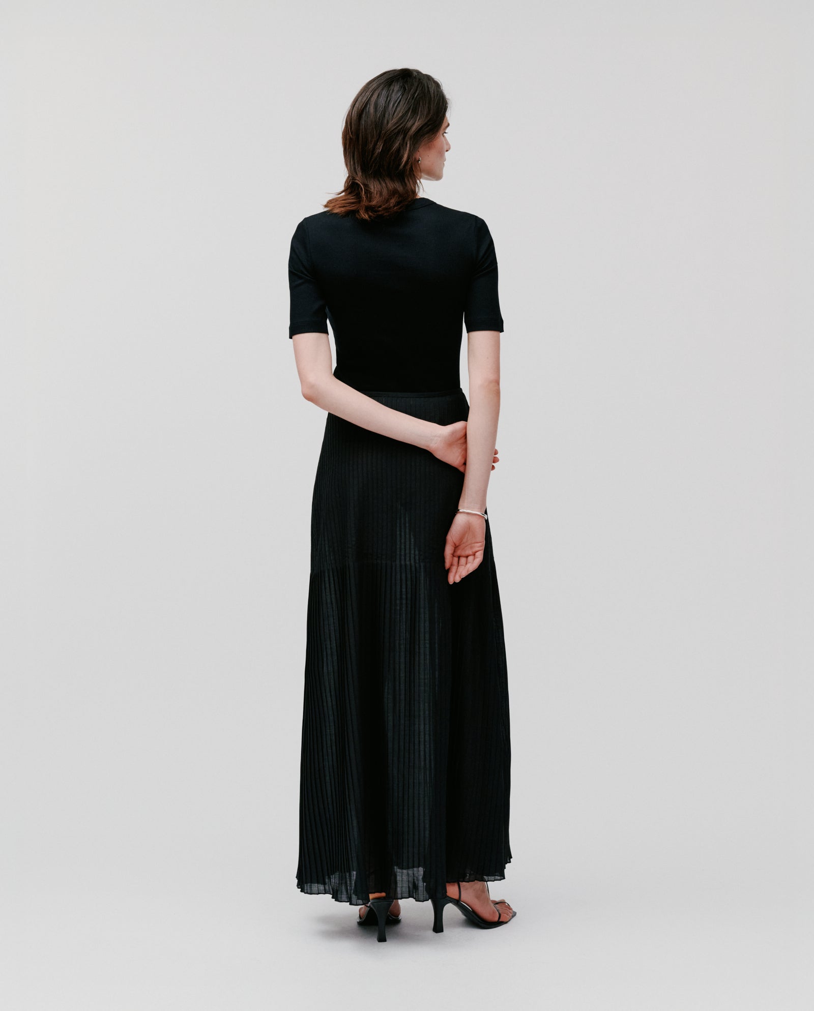 Woman wearing the IVY OAK SALENA Skirt in black plissé stands in heels, hands behind her back, facing away from the camera.