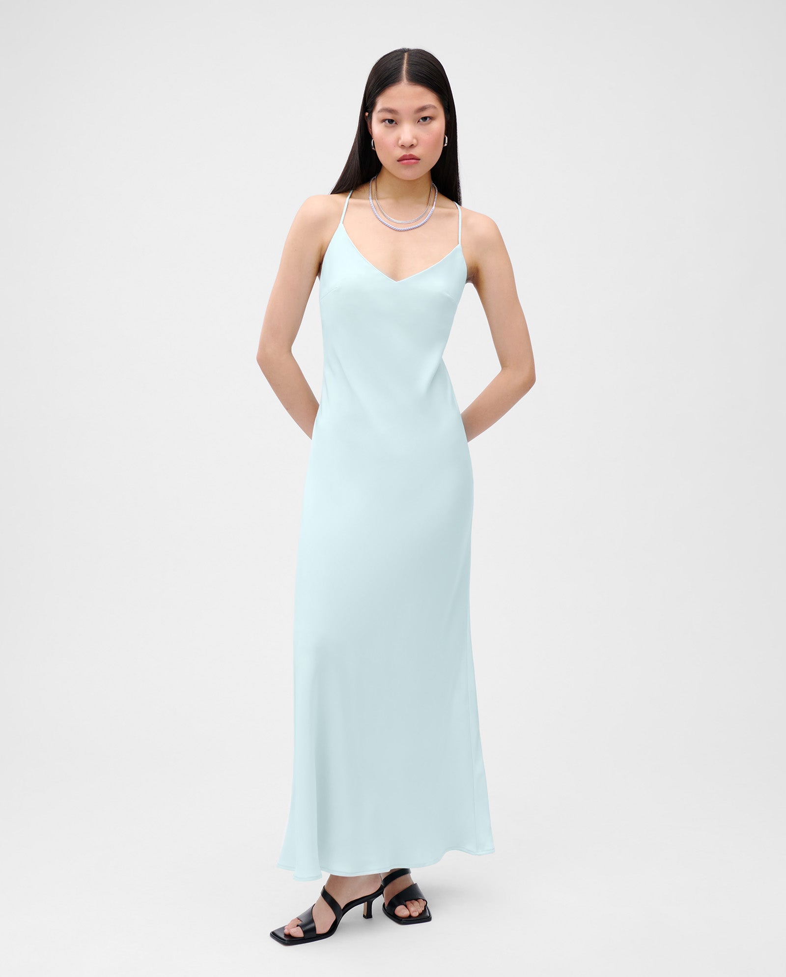 Woman in a light blue, ankle-length NOEL Dress by IVY OAK, made from shiny satin, styled with black heeled sandals and posed against a plain background.