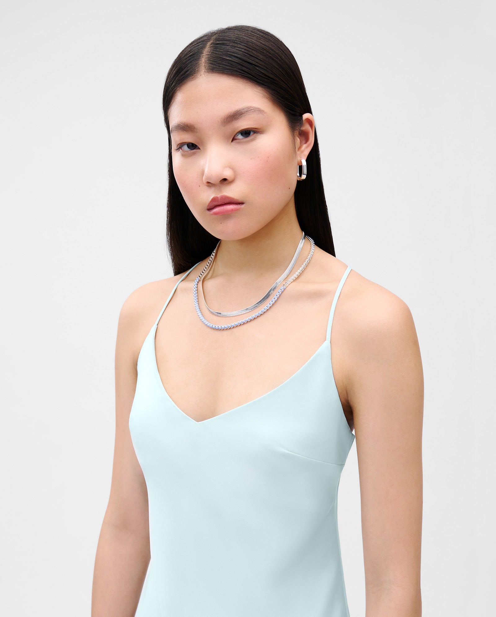 Woman with straight dark hair wears a shiny pale blue NOEL Dress by IVY OAK and layered silver necklaces, standing against a plain background.