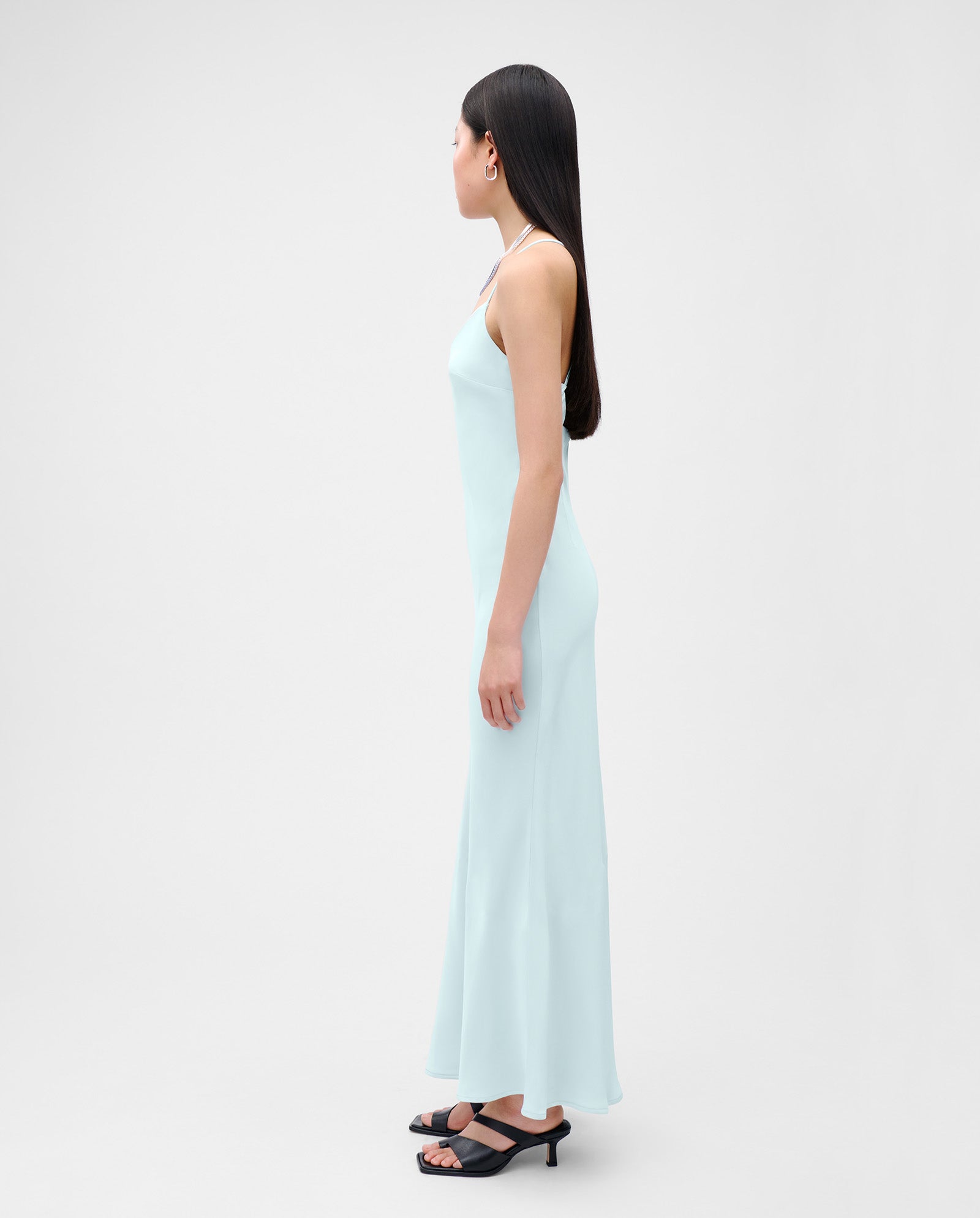 A woman in an ankle-length, light blue satin NOEL Dress by IVY OAK and black sandals stands in profile against a plain background.