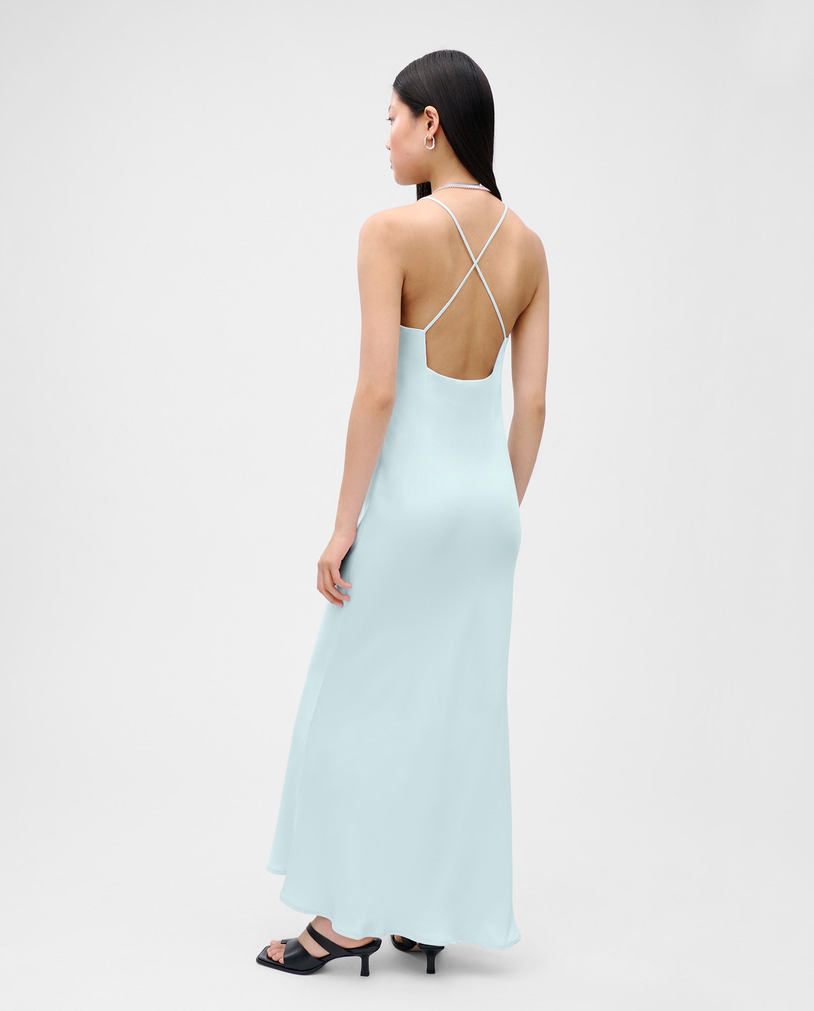 A woman wears the IVY OAK NOEL Dress—a light blue, shiny satin, backless, strappy ankle-length slipdress—with black heels, standing against a plain background.