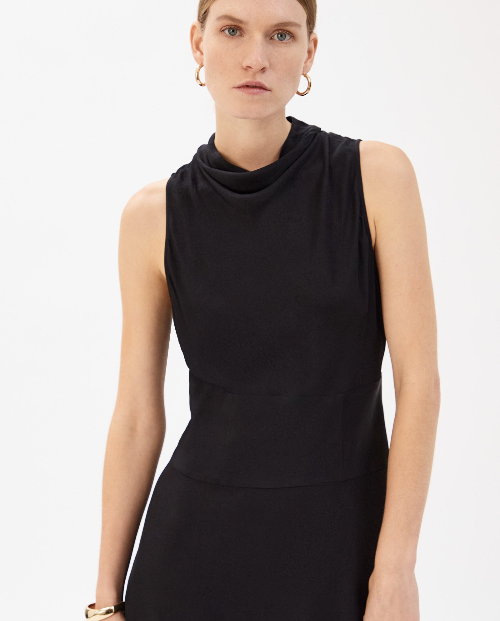 A woman wearing a sleeveless black NABINA Dress by IVY OAK with a waterfall collar stands against a plain white background, looking at the camera.