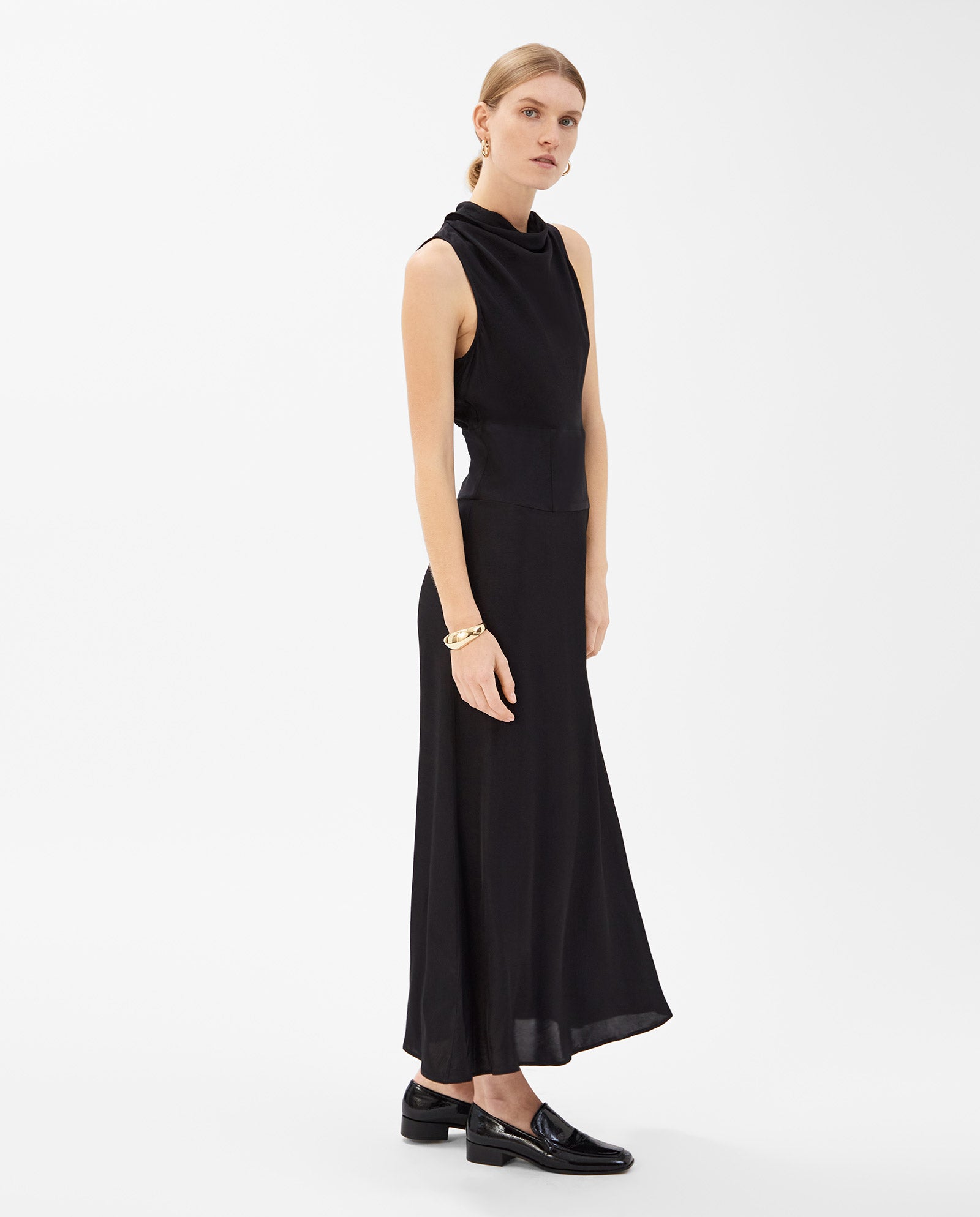 A woman wears a sleeveless black IVY OAK NABINA dress with a waterfall collar and black loafers, standing against a plain white background.