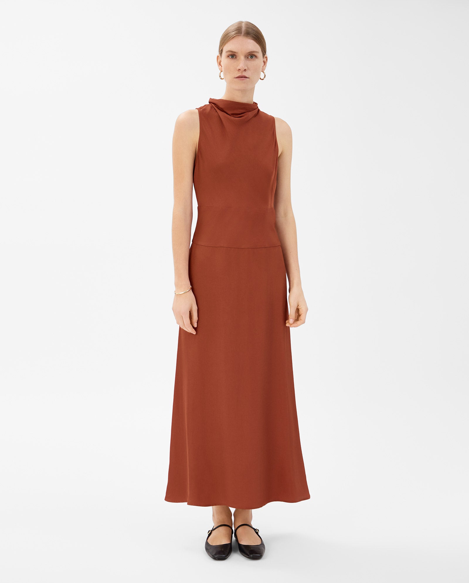 A woman wears the sleeveless NABINA Dress by IVY OAK in rust, featuring a waterfall collar and crafted from sustainable acetate, styled with black flats against a white backdrop.