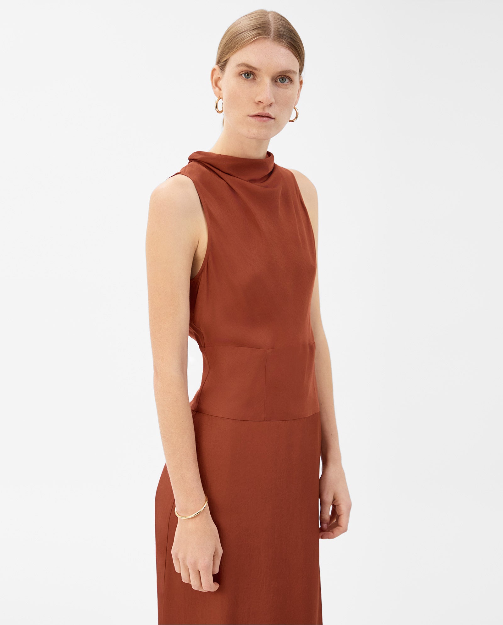 A woman models the IVY OAK NABINA Dress, a sleeveless rust-hued style with a waterfall collar, paired with gold hoop earrings against a white background.