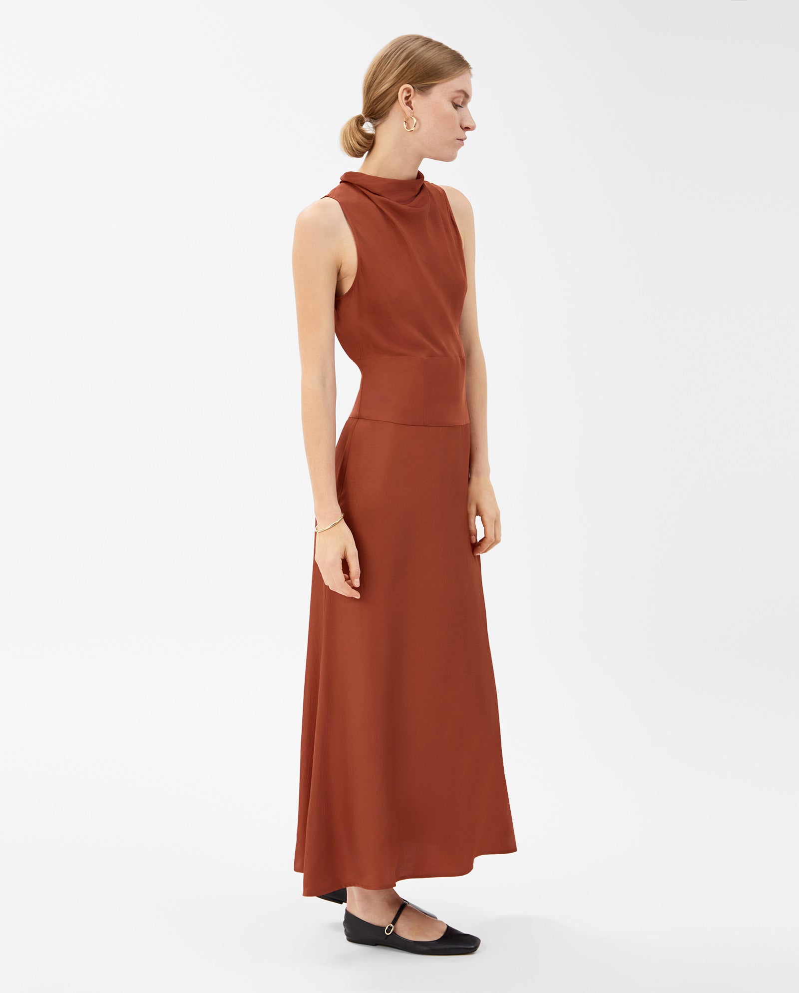 A woman stands sideways in a sleeveless rust NABINA Dress by IVY OAK, featuring a waterfall collar and crafted from sustainable acetate, paired with black flats.