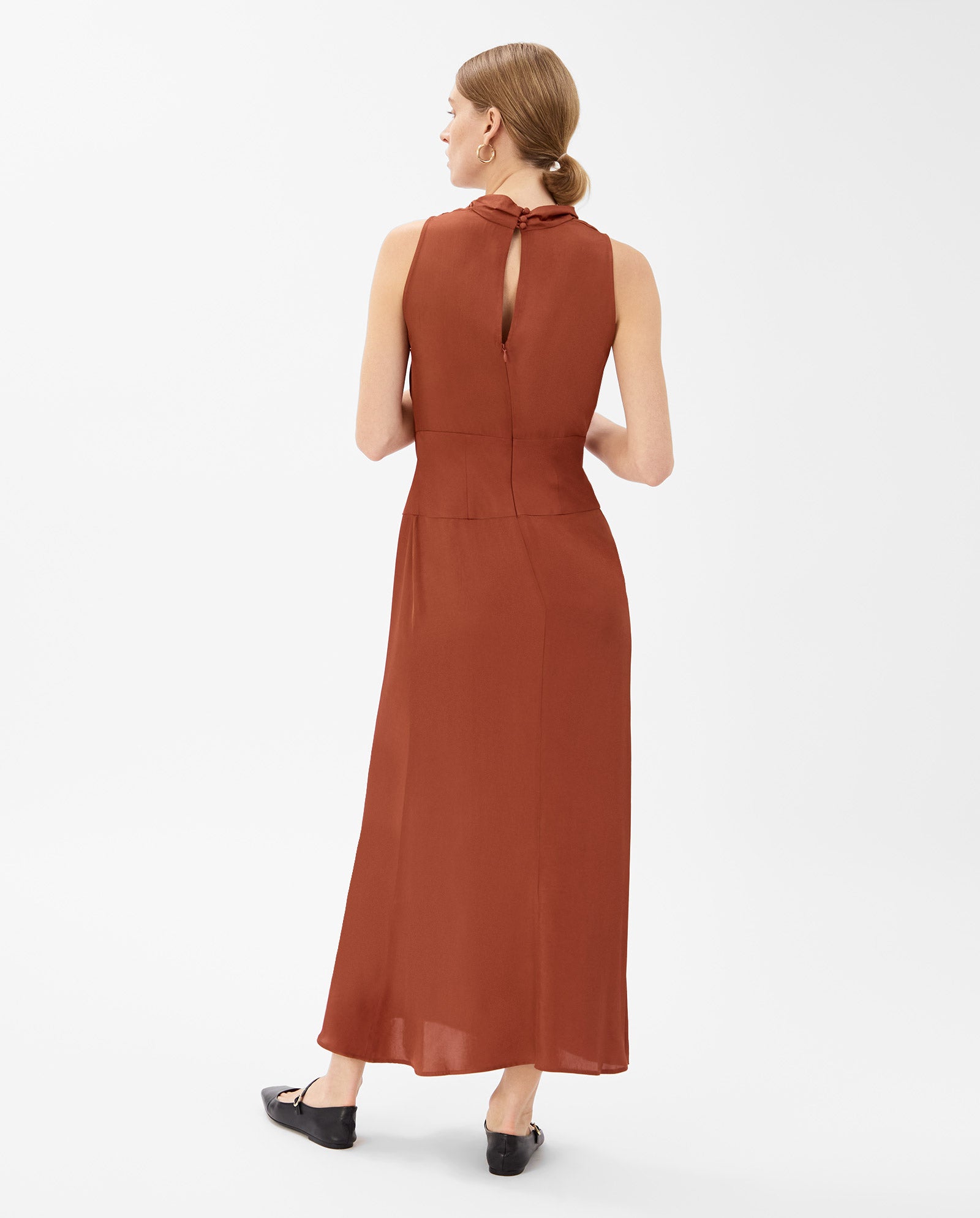 A woman stands facing away in a sleeveless rust NABINA Dress by IVY OAK, crafted from sustainable acetate with a waterfall collar and back keyhole detail.