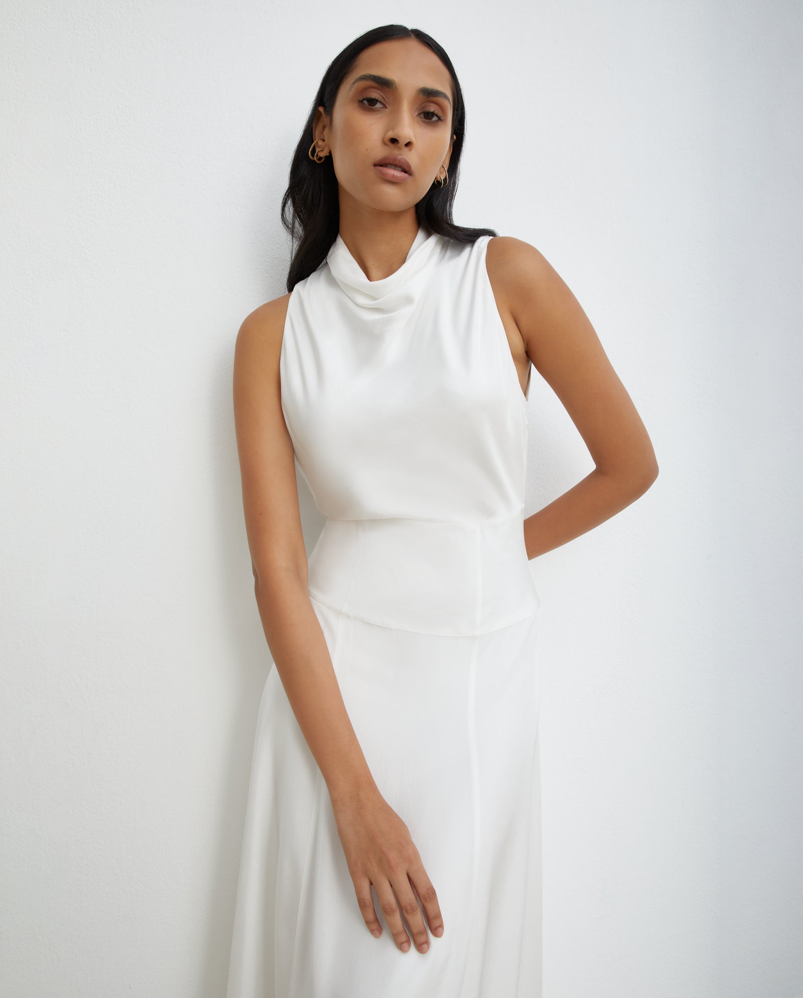 A woman in the IVY OAK NABINA LOU Bridaldress, a sleeveless Naia acetate A-line wedding gown, stands against a plain white wall with one arm resting across her waist.