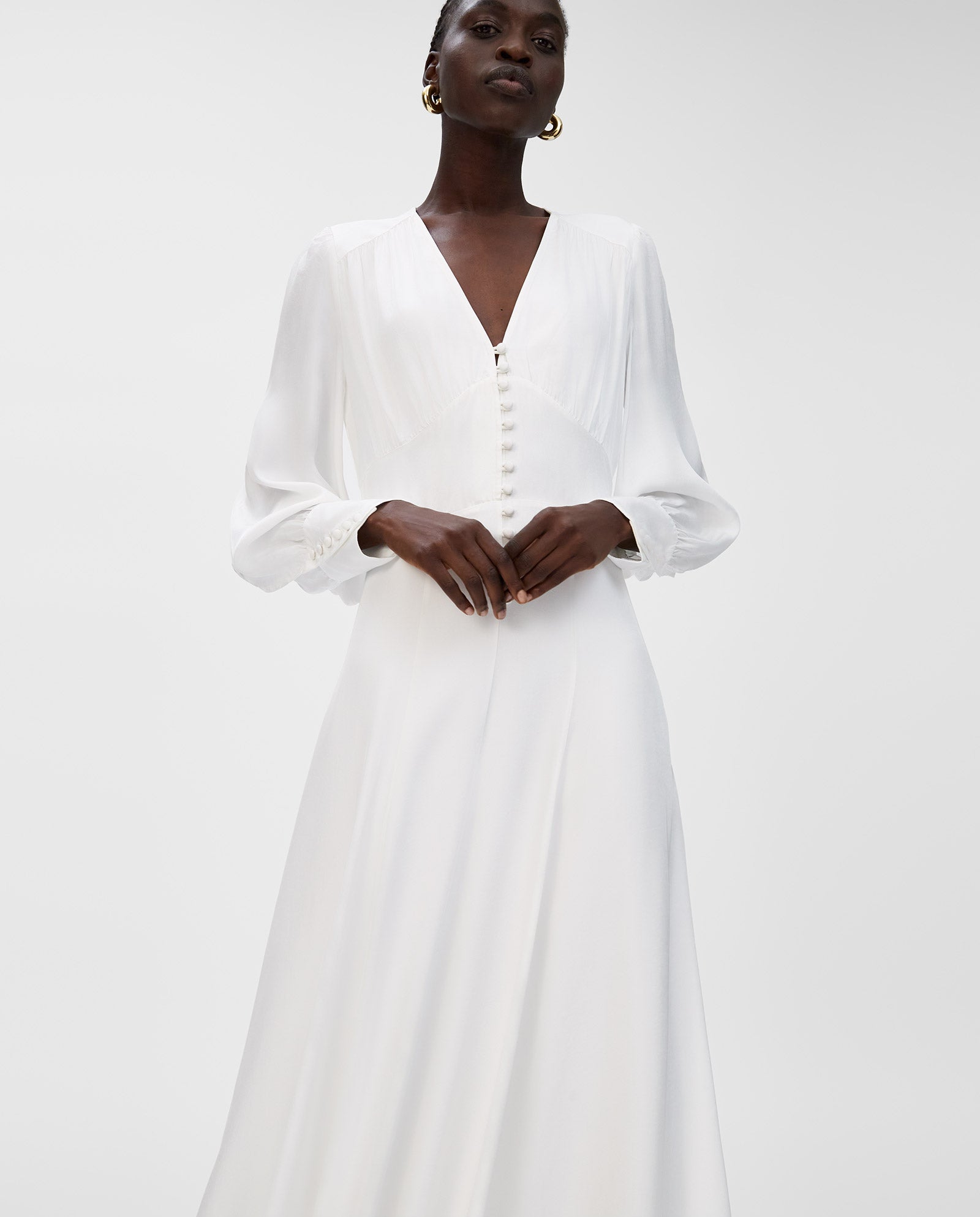 A woman wears the IVY OAK NYSSA ROSE Bridaldress, a long-sleeve white dress with a deep V-neckline and front buttons, standing against a plain background.